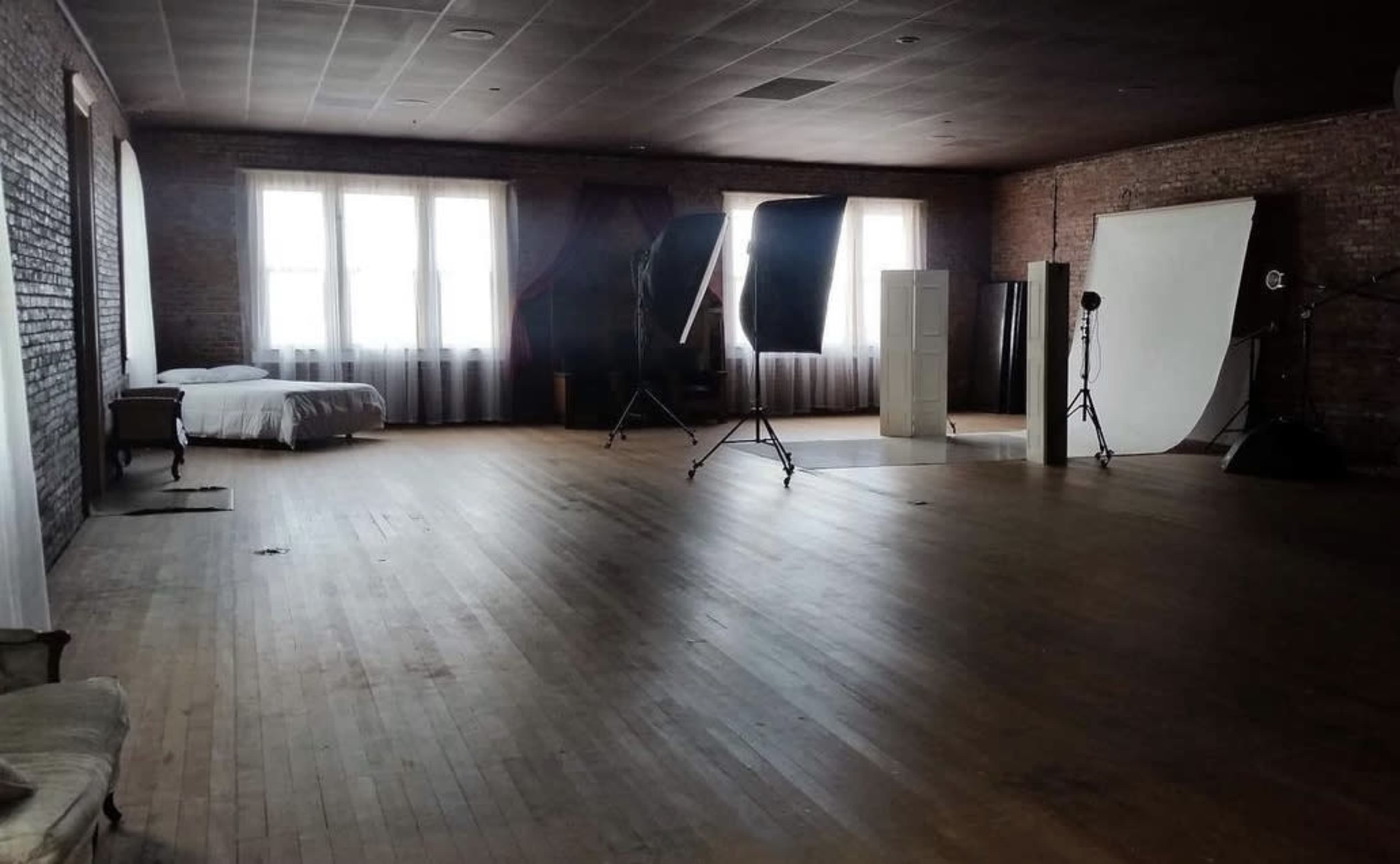 An empty studio space featuring wooden floors, large windows, a bed, and various photography equipment set up in the background.