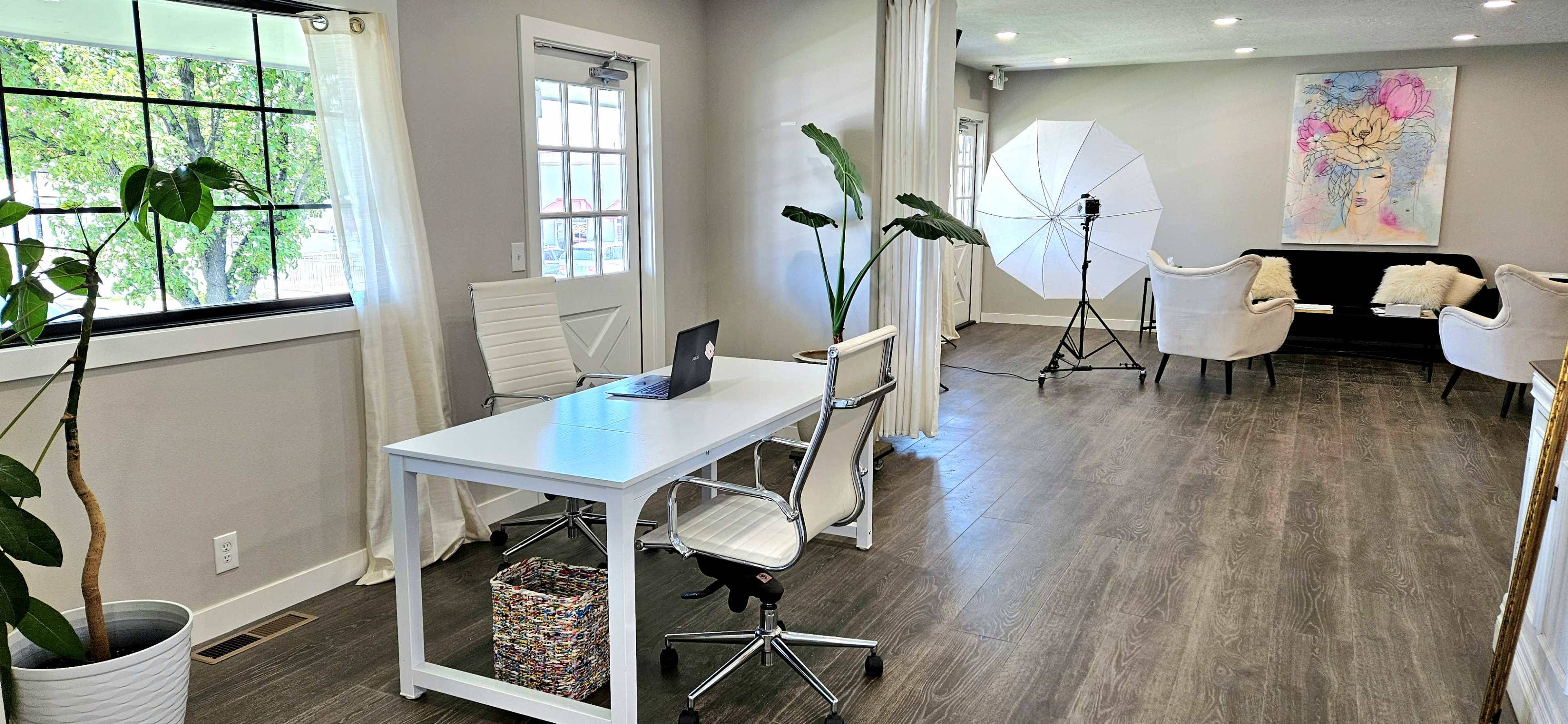 A modern office space featuring a white desk with a laptop, a large umbrella reflector for photography, and a seating area with two chairs and a decorative plant.