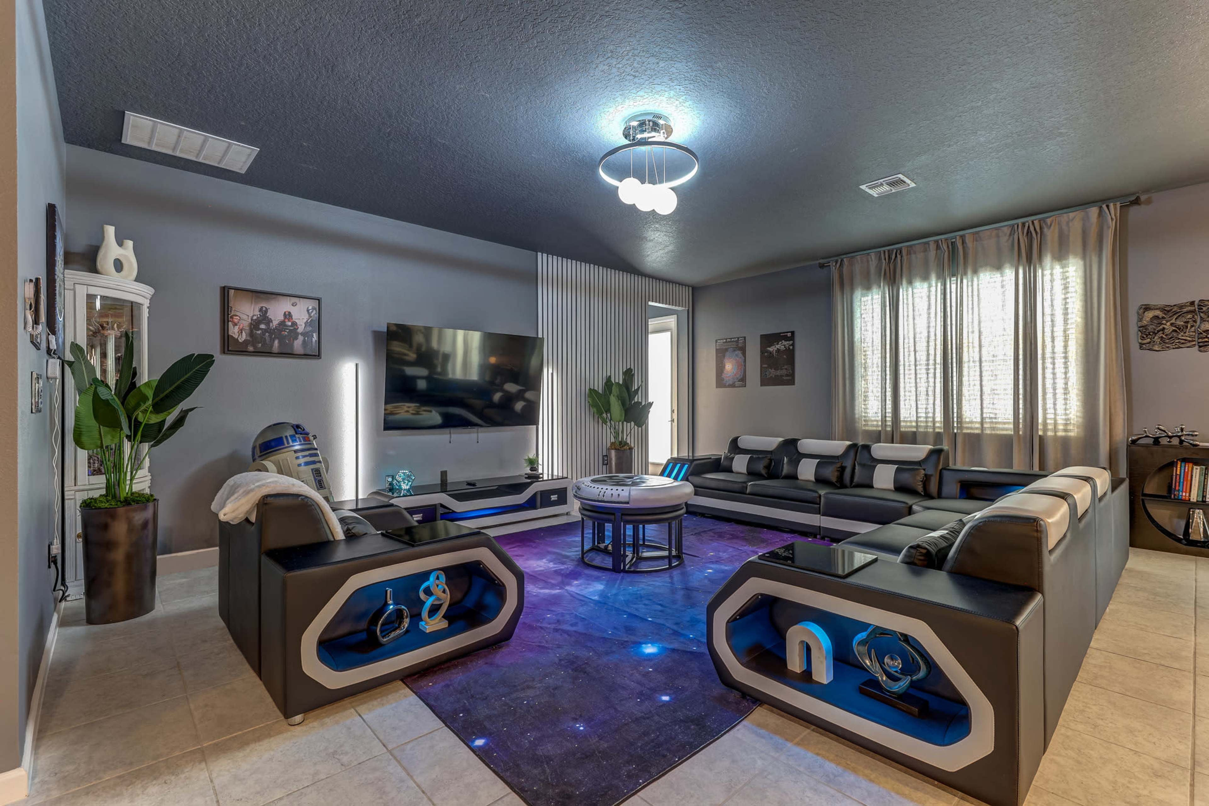 The image shows a modern living room with a spacious layout, featuring a sectional sofa, a round coffee table, and a large TV mounted on the wall, all set against a backdrop of gray walls and soft lighting.