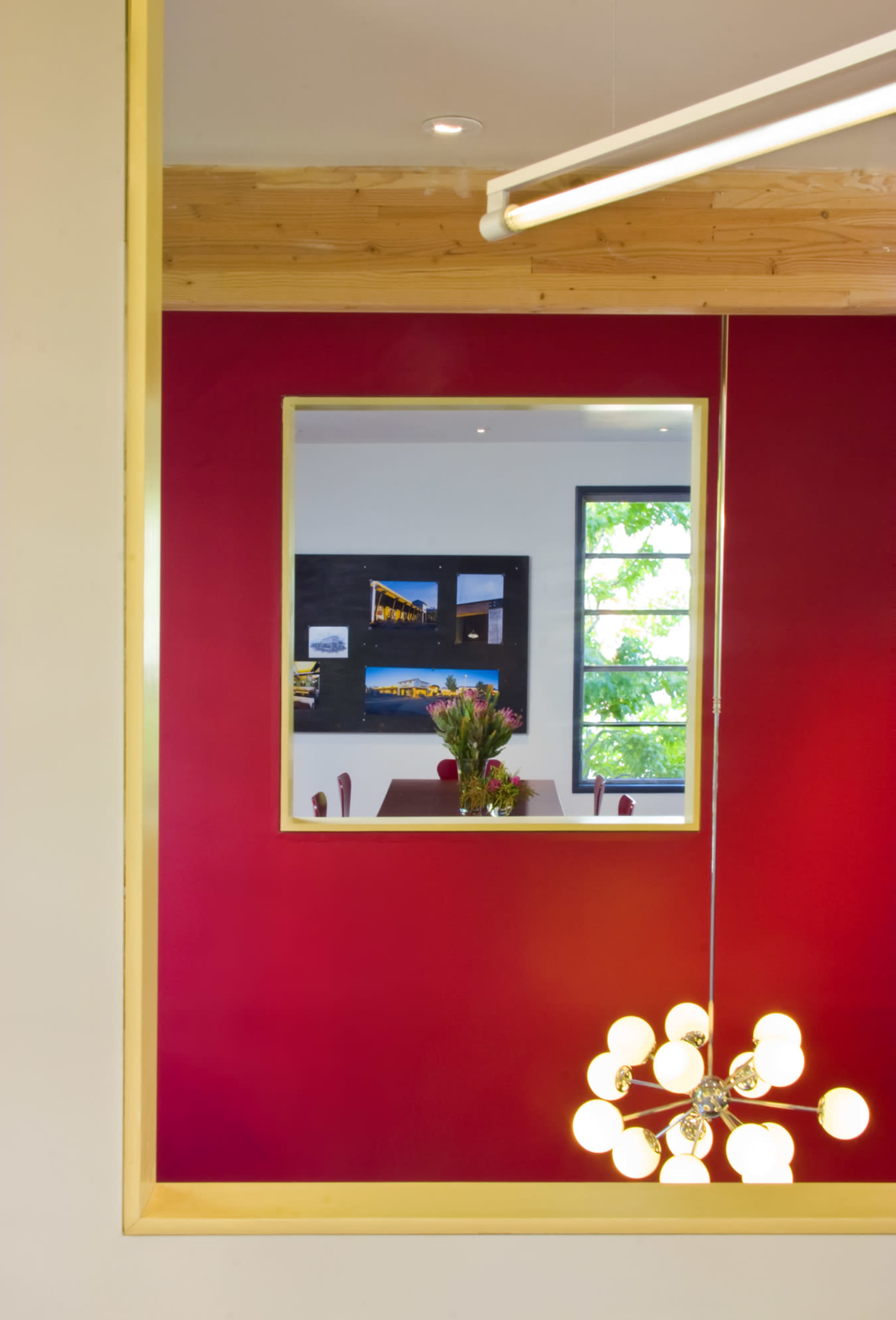 A rectangular mirror is framed in gold against a red wall, reflecting a dining area with a table and flower vase in the background.