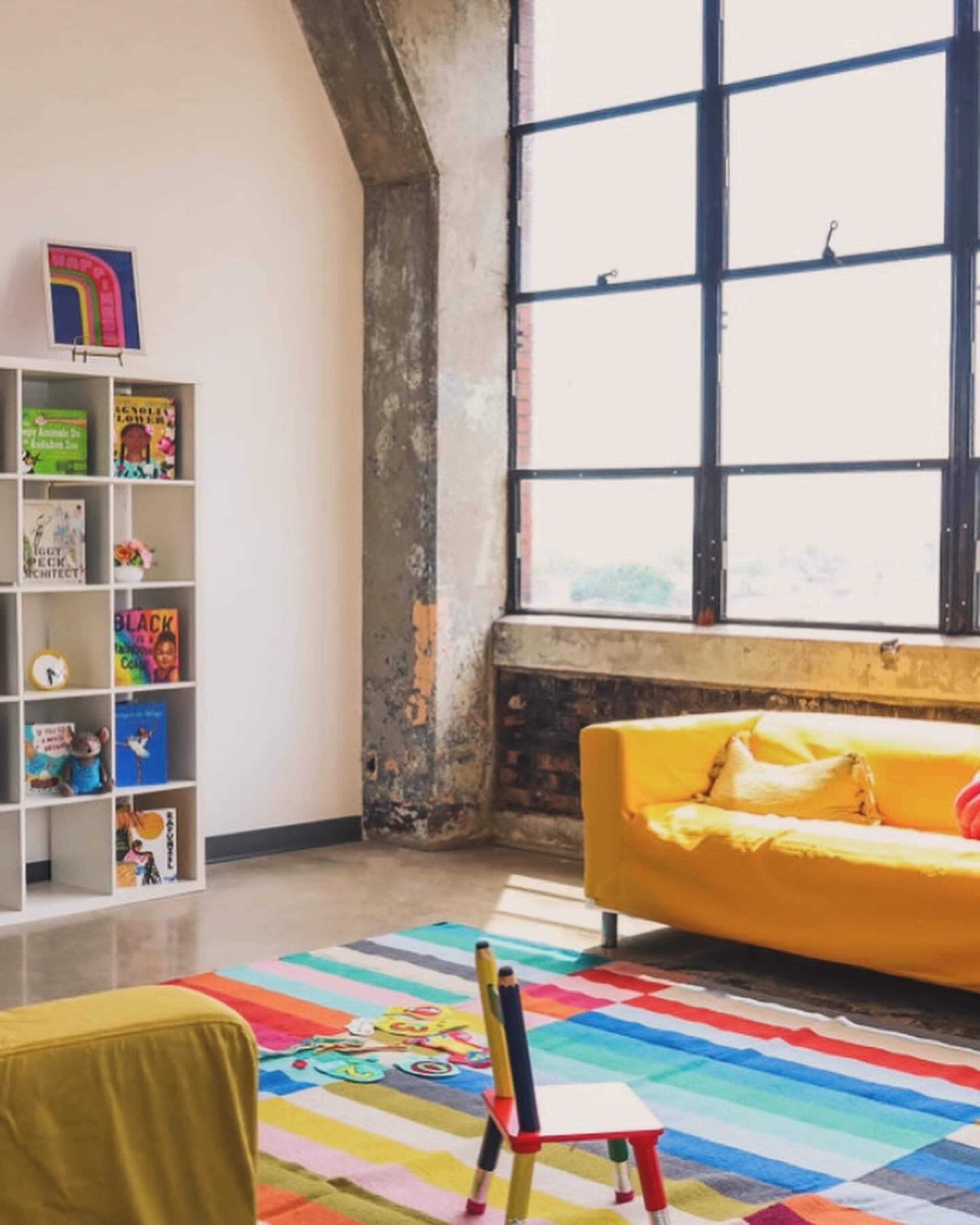 A bright, modern living space features a yellow couch, a colorful rug, and a bookshelf filled with children's books beside large windows.