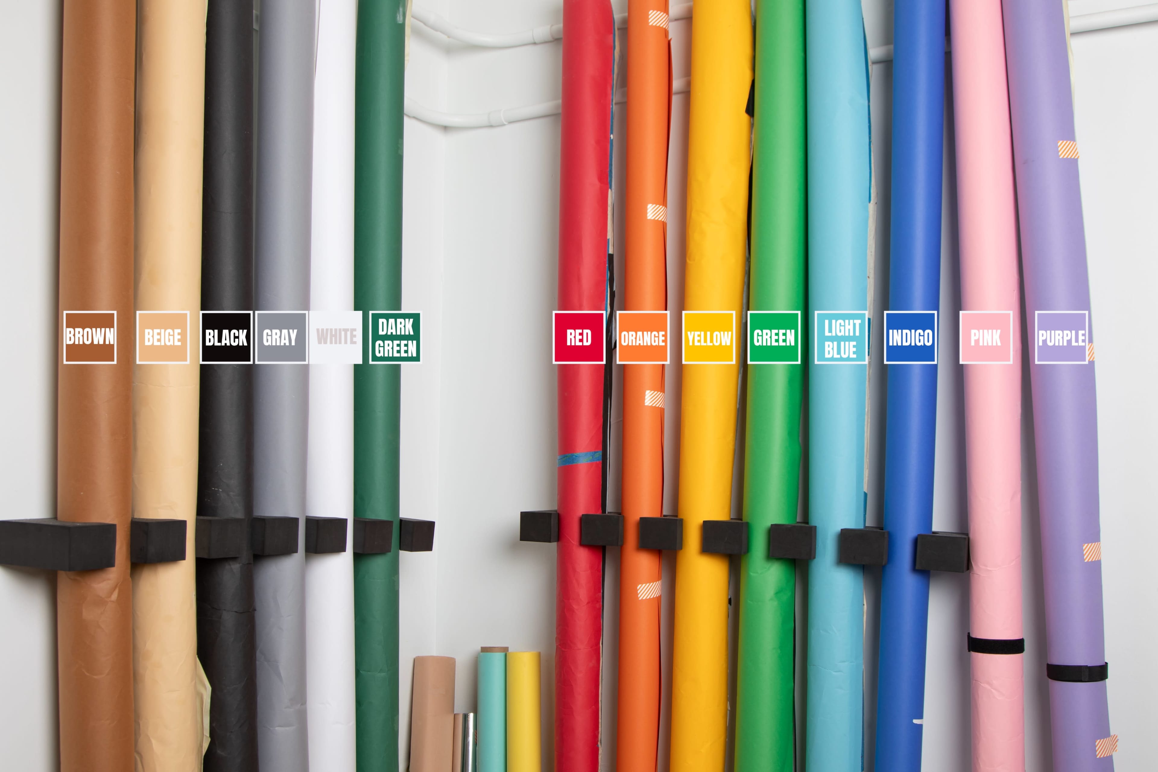 The image shows a neatly organized collection of rolls of colored paper or vinyl stored vertically on a wall, labeled with their respective colors.