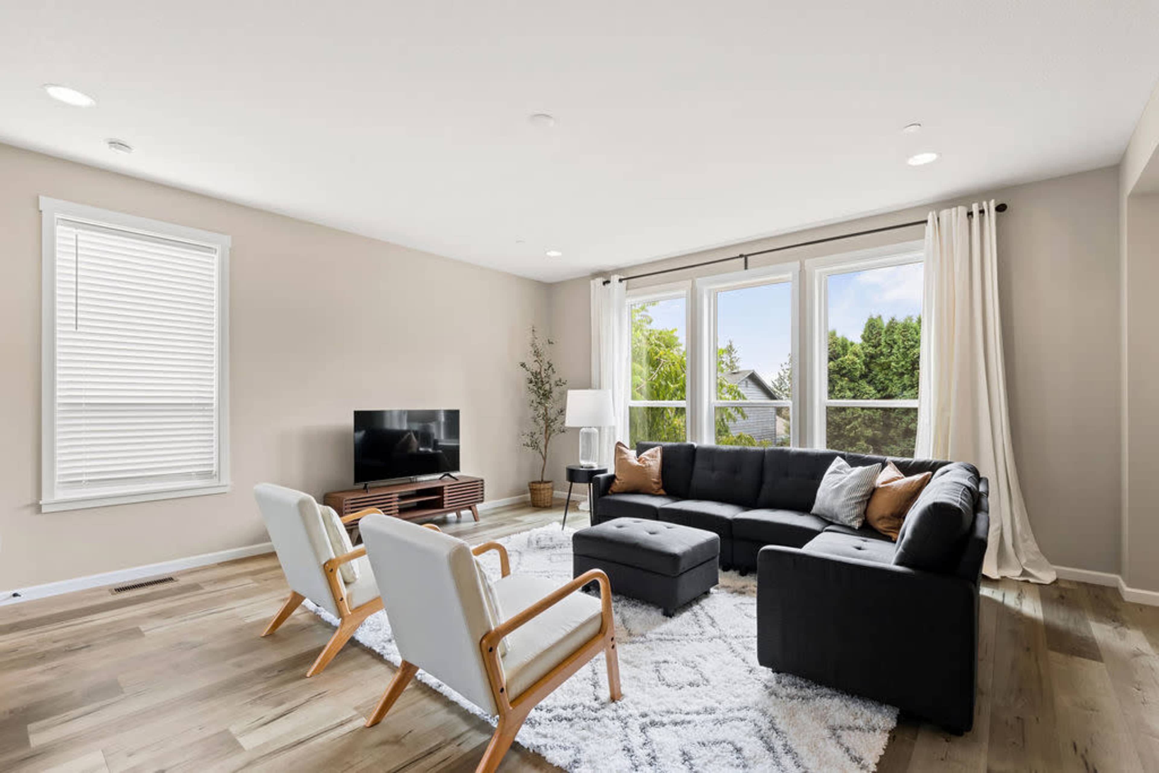 A modern living room features a black sectional sofa, two white chairs, a television on a wooden stand, and large windows overlooking greenery.