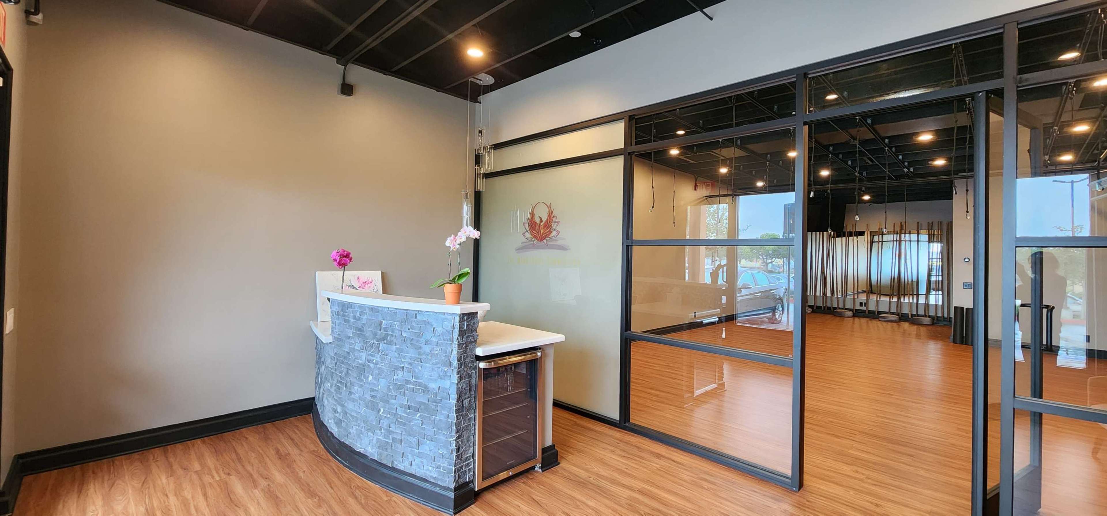 A modern reception area with a stone front desk, a potted orchid, and large glass panels leading into a spacious room with wooden flooring.