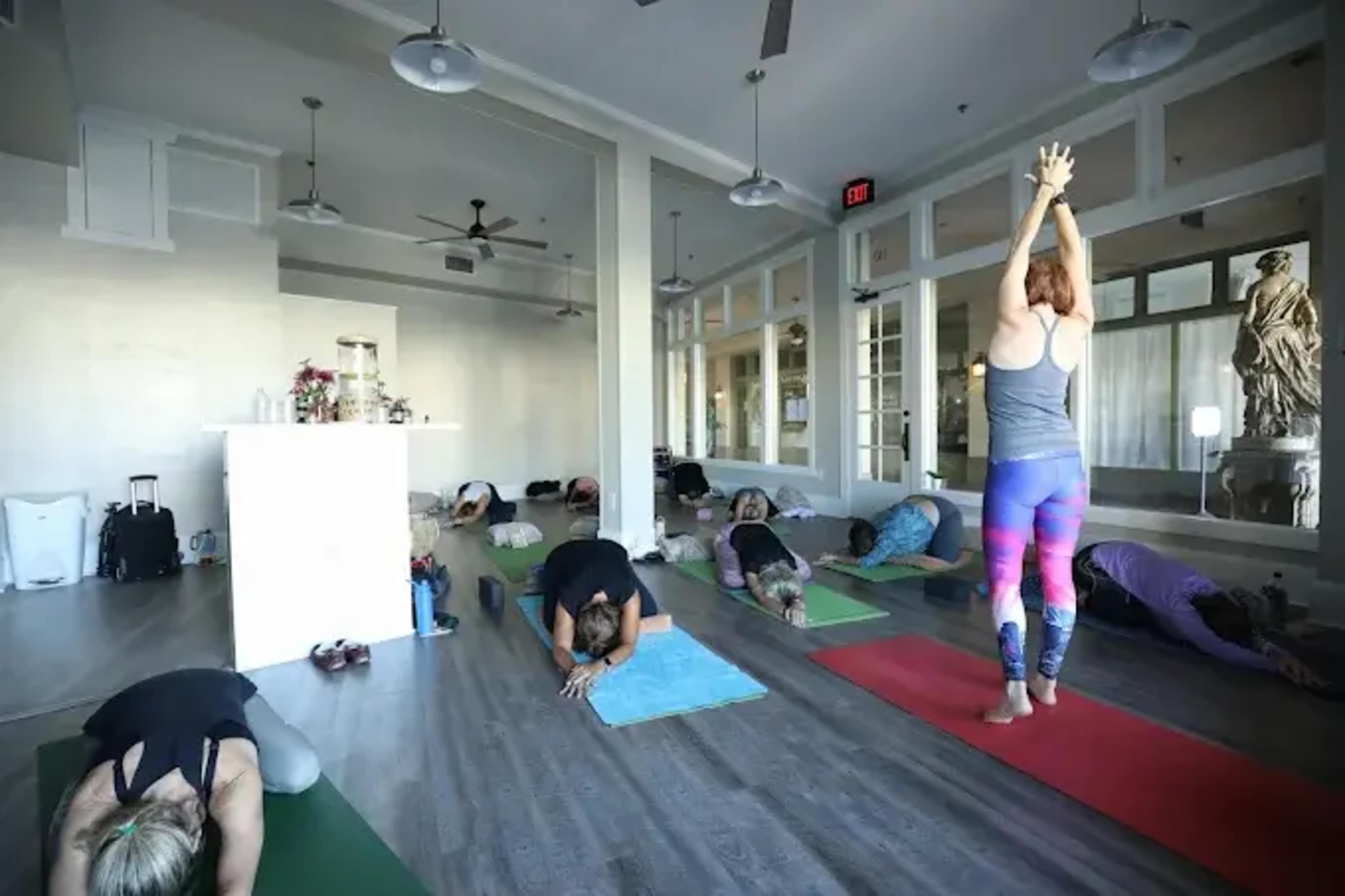 A group of individuals is practicing yoga in a studio with large windows and wooden floors, while one person stands at the front performing a pose.