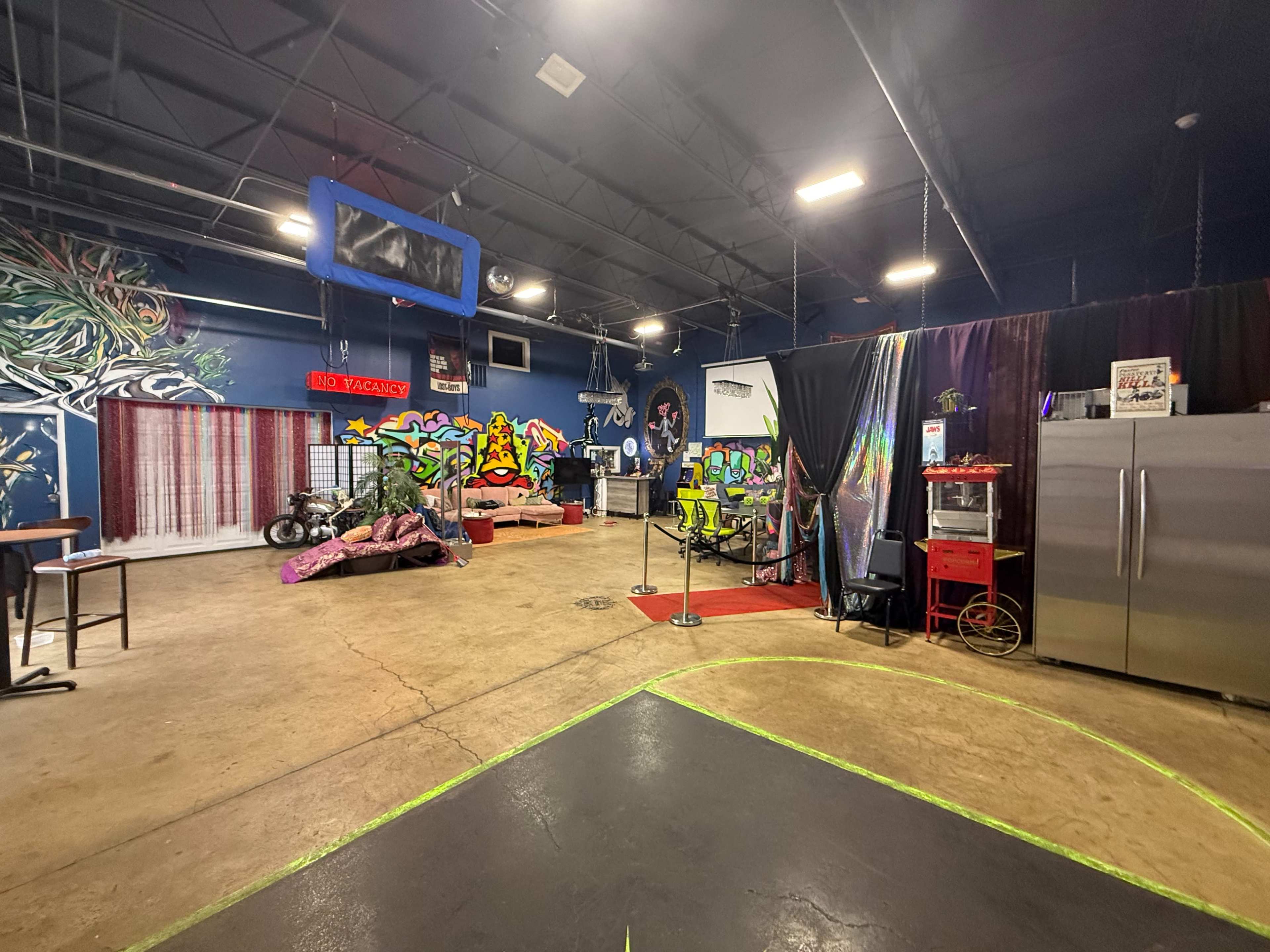 The image shows an eclectic indoor space featuring colorful graffiti, a seating area with couches, various objects scattered around, and a stainless steel refrigerator in one corner.