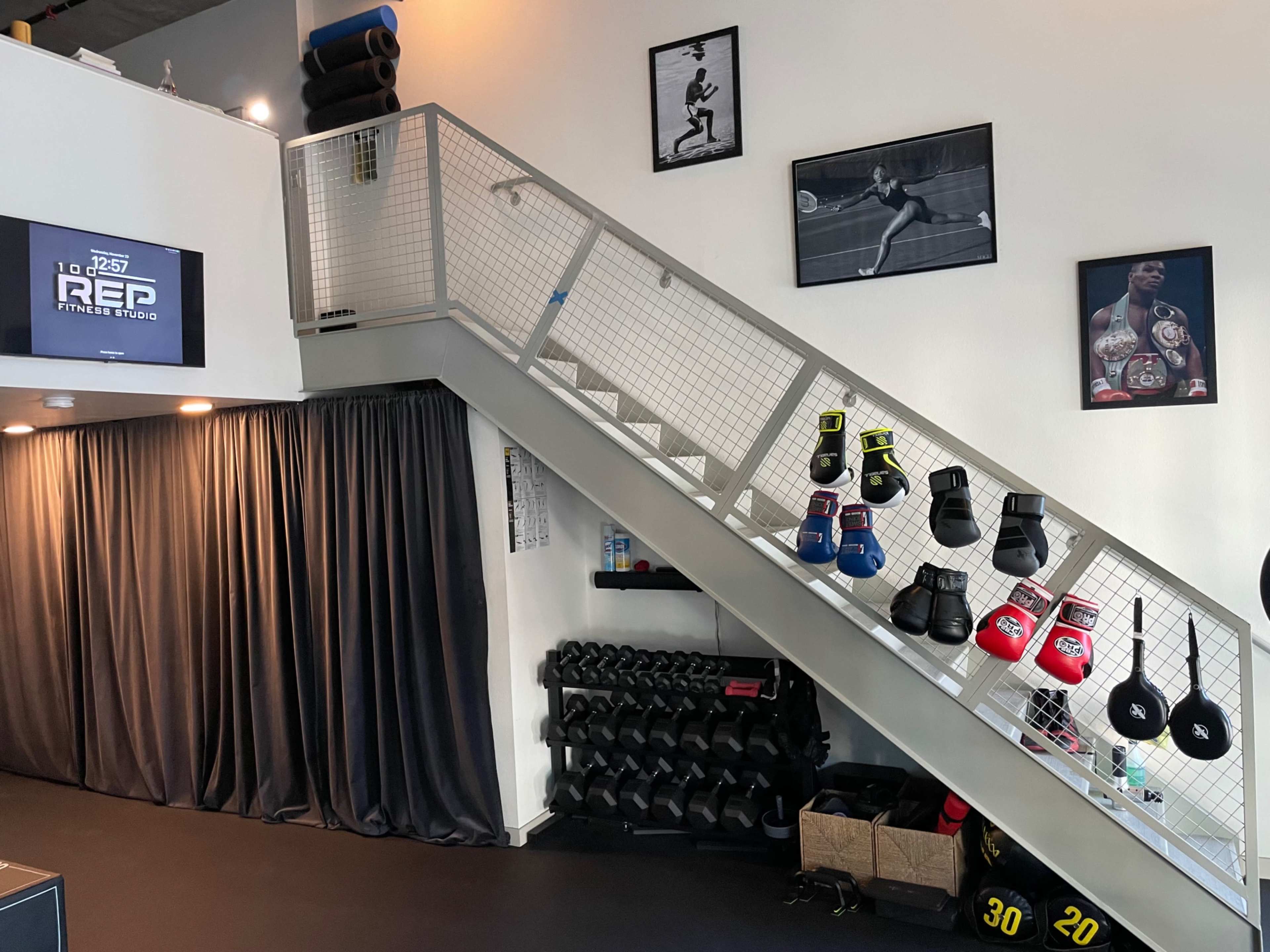The image shows an interior gym space featuring a staircase with boxing gloves hanging on the railing, dumbbells on the floor, and framed photographs on the walls.