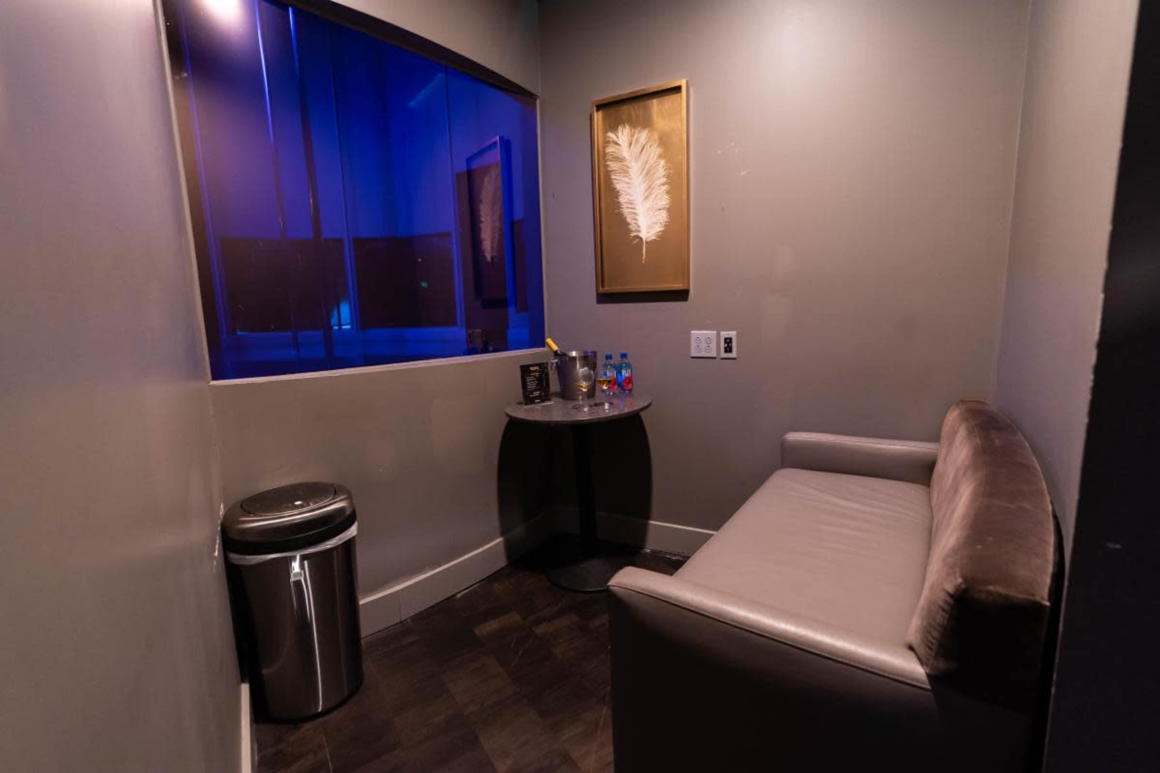 A small room with a gray couch, a round table with colorful objects, a trash can, and a large window with blue lighting.