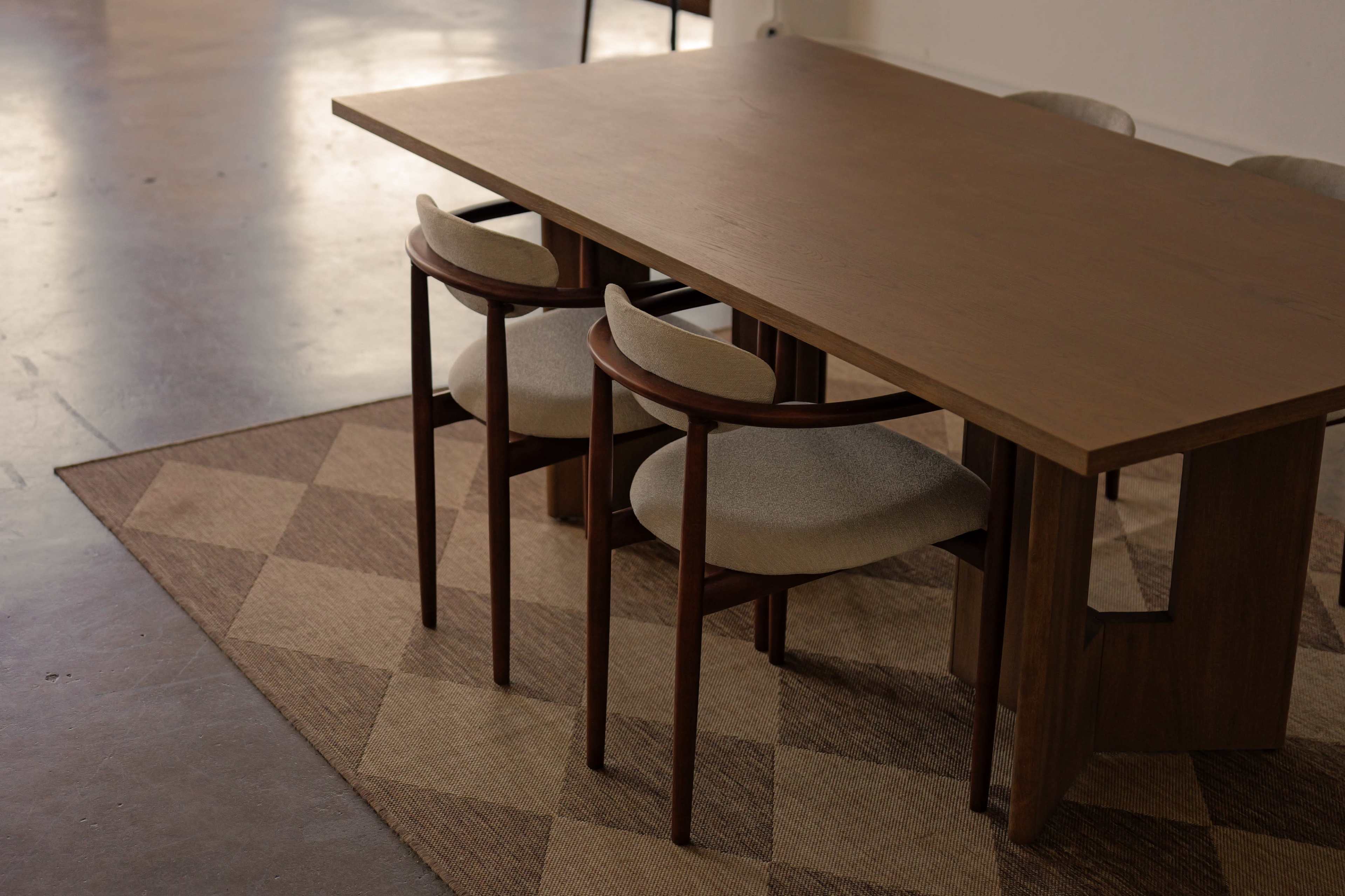 A wooden table with a rectangular top is accompanied by two upholstered chairs, all situated on a patterned rug in an indoor space.
