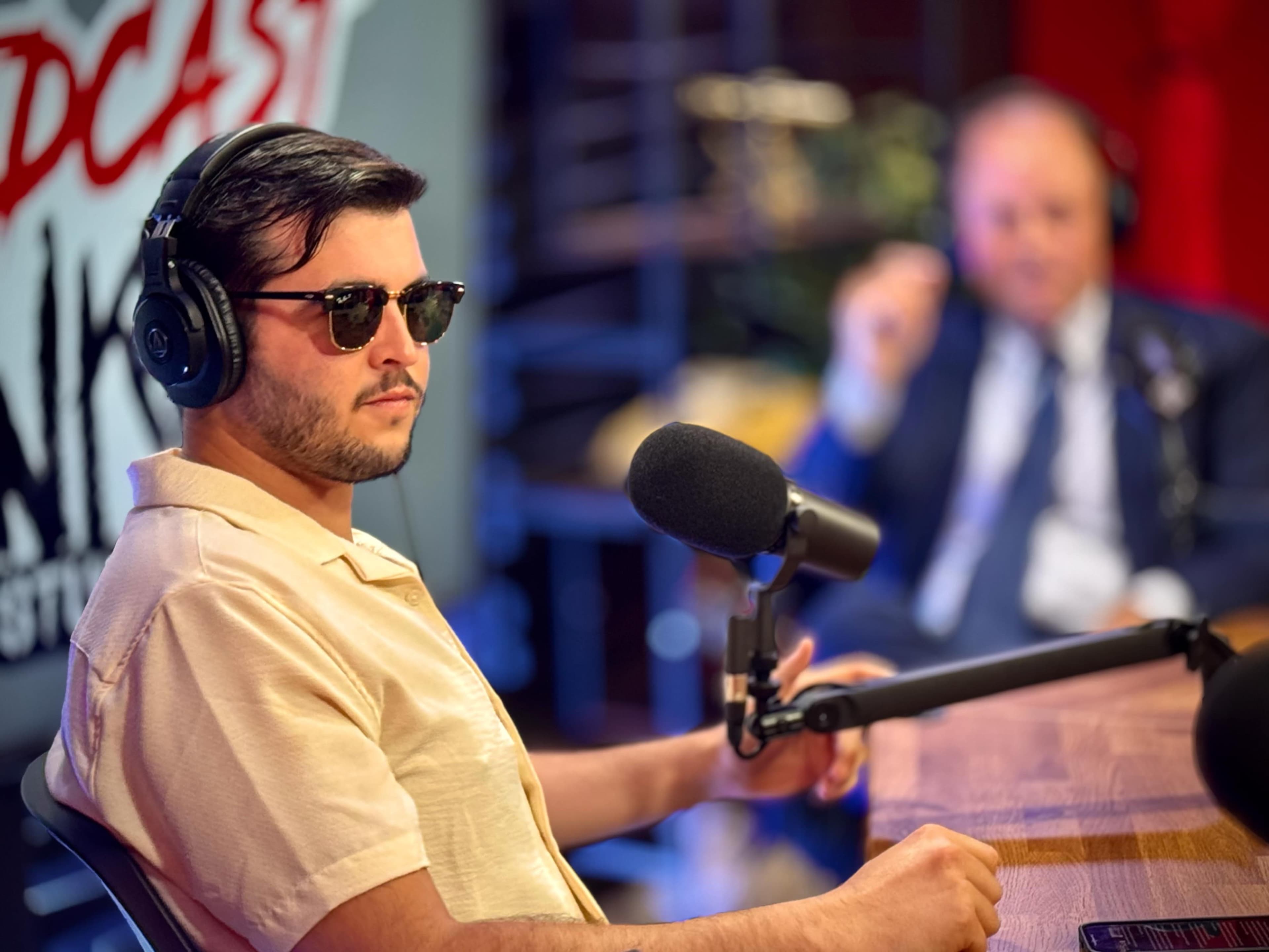 A man wearing sunglasses sits at a podcast table with a microphone, while a second individual is partially visible in the background.