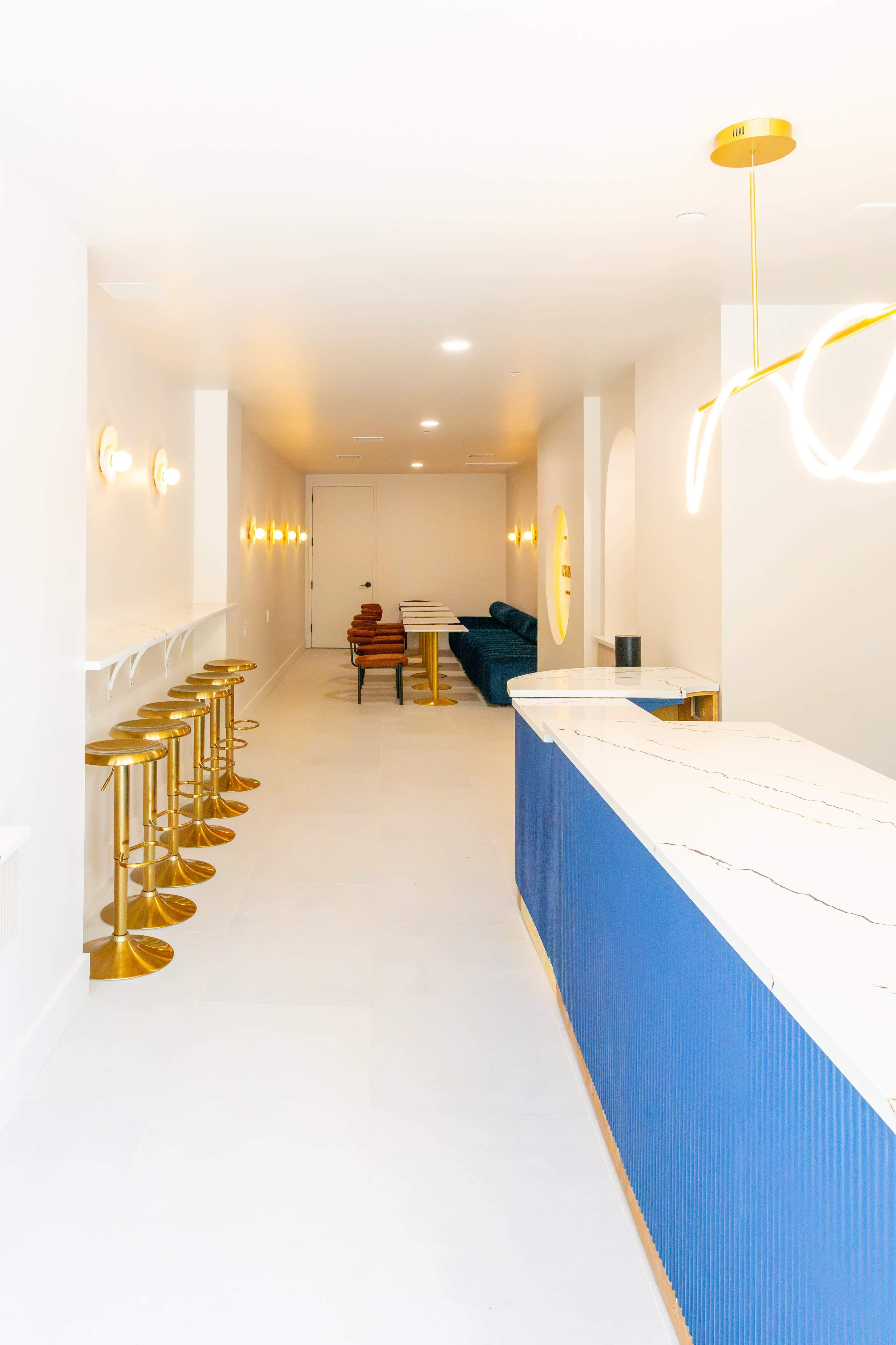 The image shows a modern interior space with a long bar featuring gold stools, blue cabinetry, and a row of light fixtures along the walls.