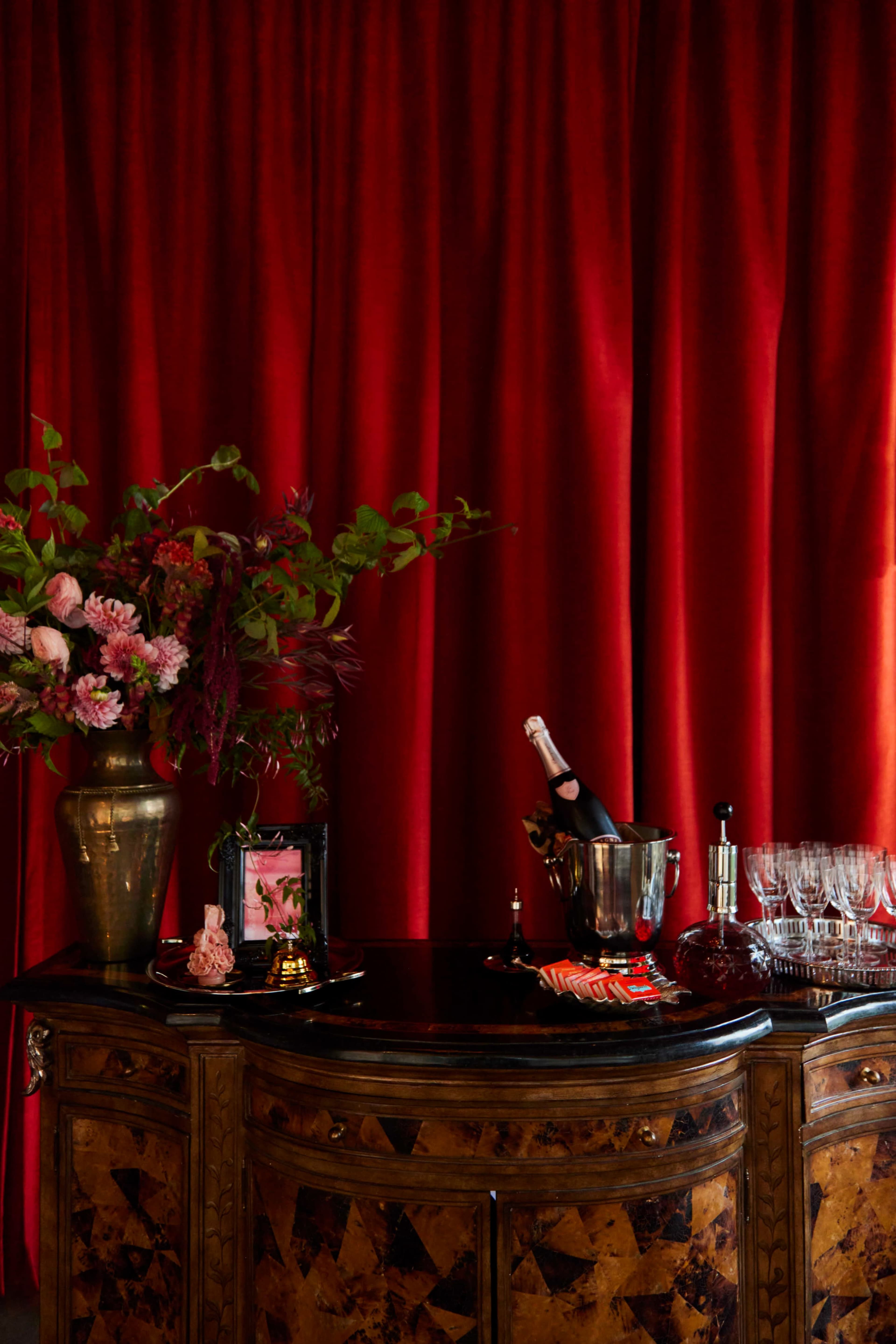 A vintage wooden sideboard displays a bucket of champagne, glassware, and a floral arrangement against a backdrop of deep red curtains.