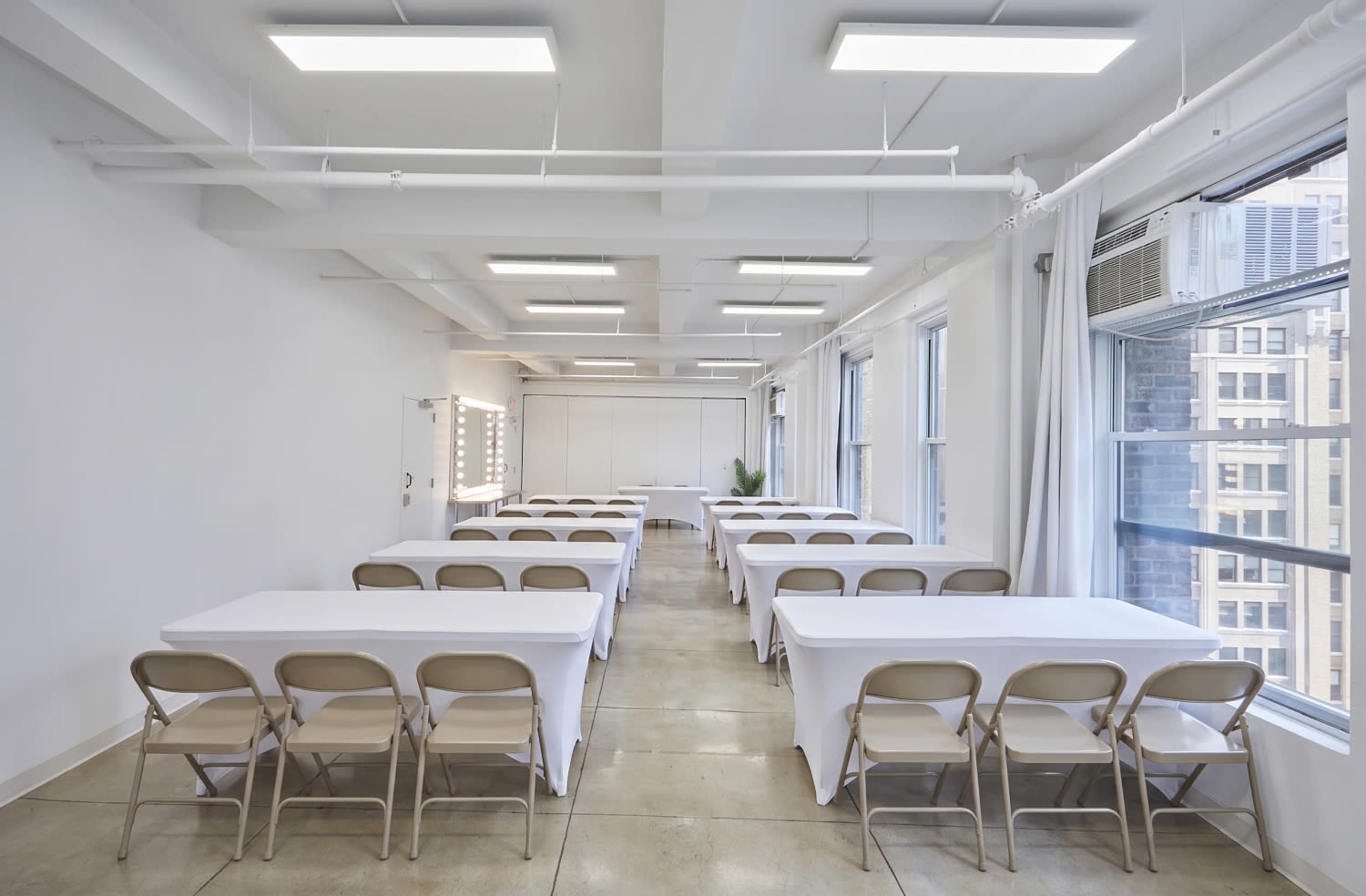 The image shows a well-lit meeting room with several tables arranged in rows, covered with white tablecloths, and large windows letting in natural light.
