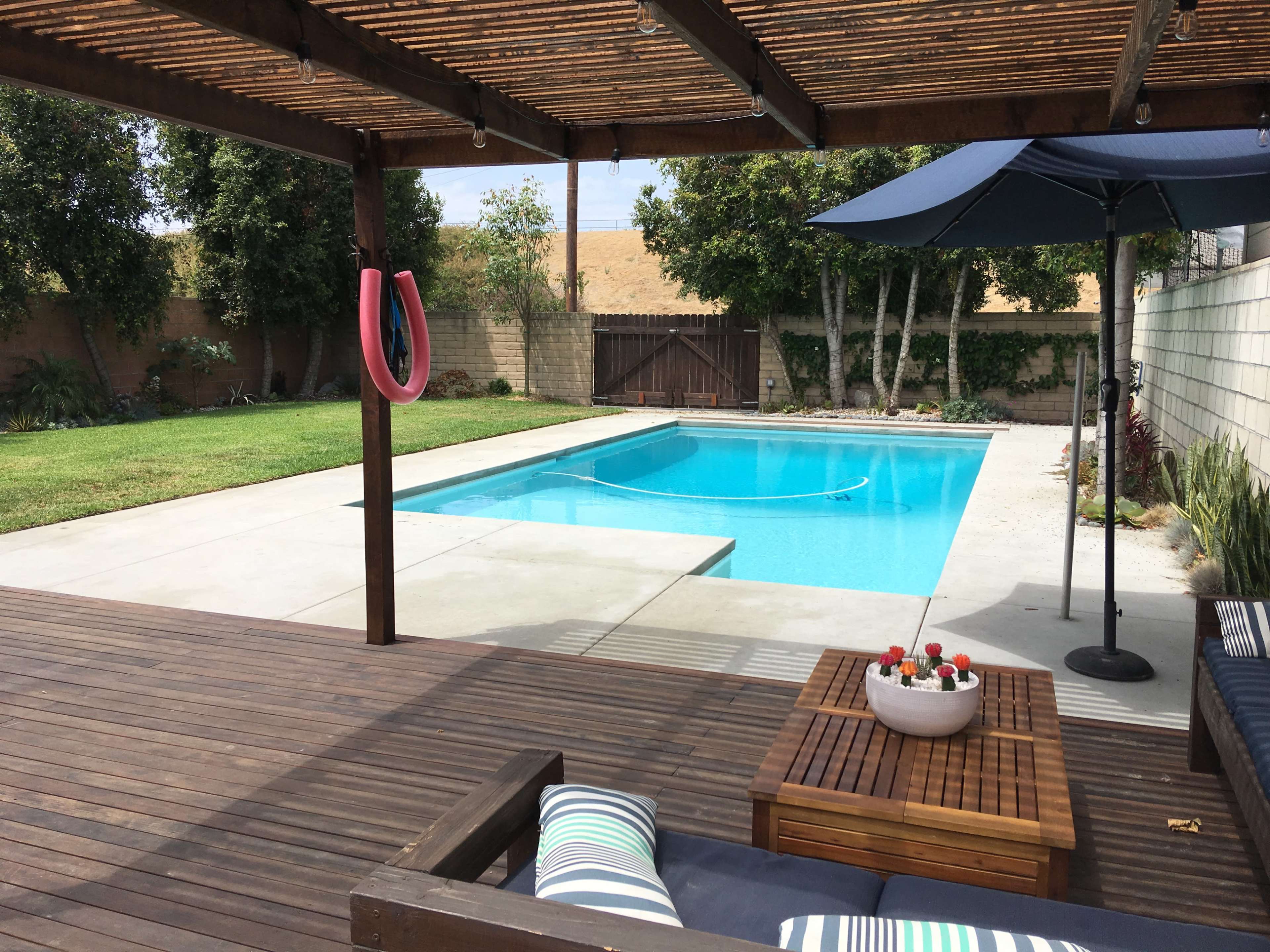 A well-maintained backyard features a swimming pool surrounded by a patio, lounge furniture, and a covered area with a hanging ring.