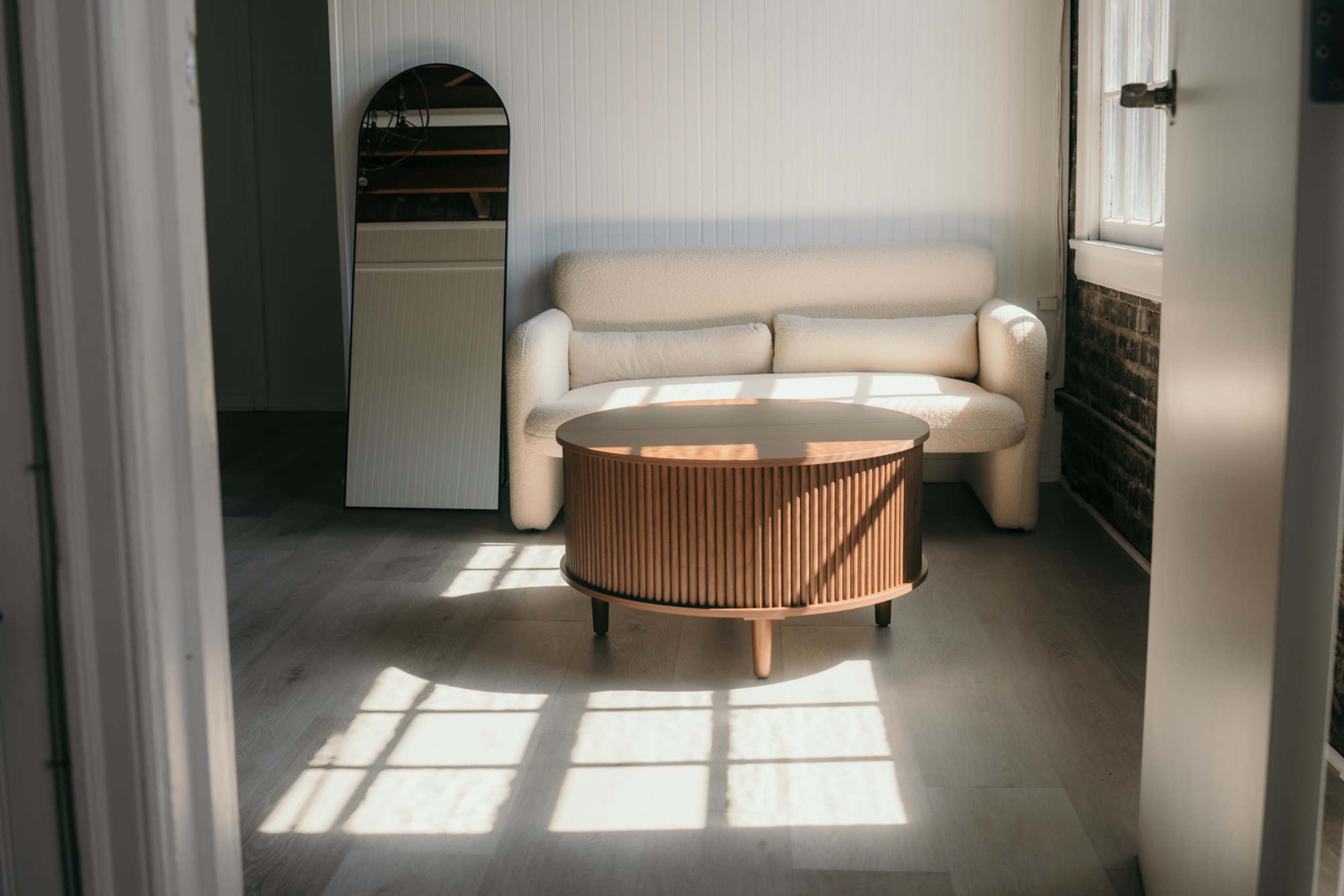 The image shows a minimalist living space featuring a light-colored sofa, a round coffee table with vertical wood slats, and a mirror reflecting natural light.