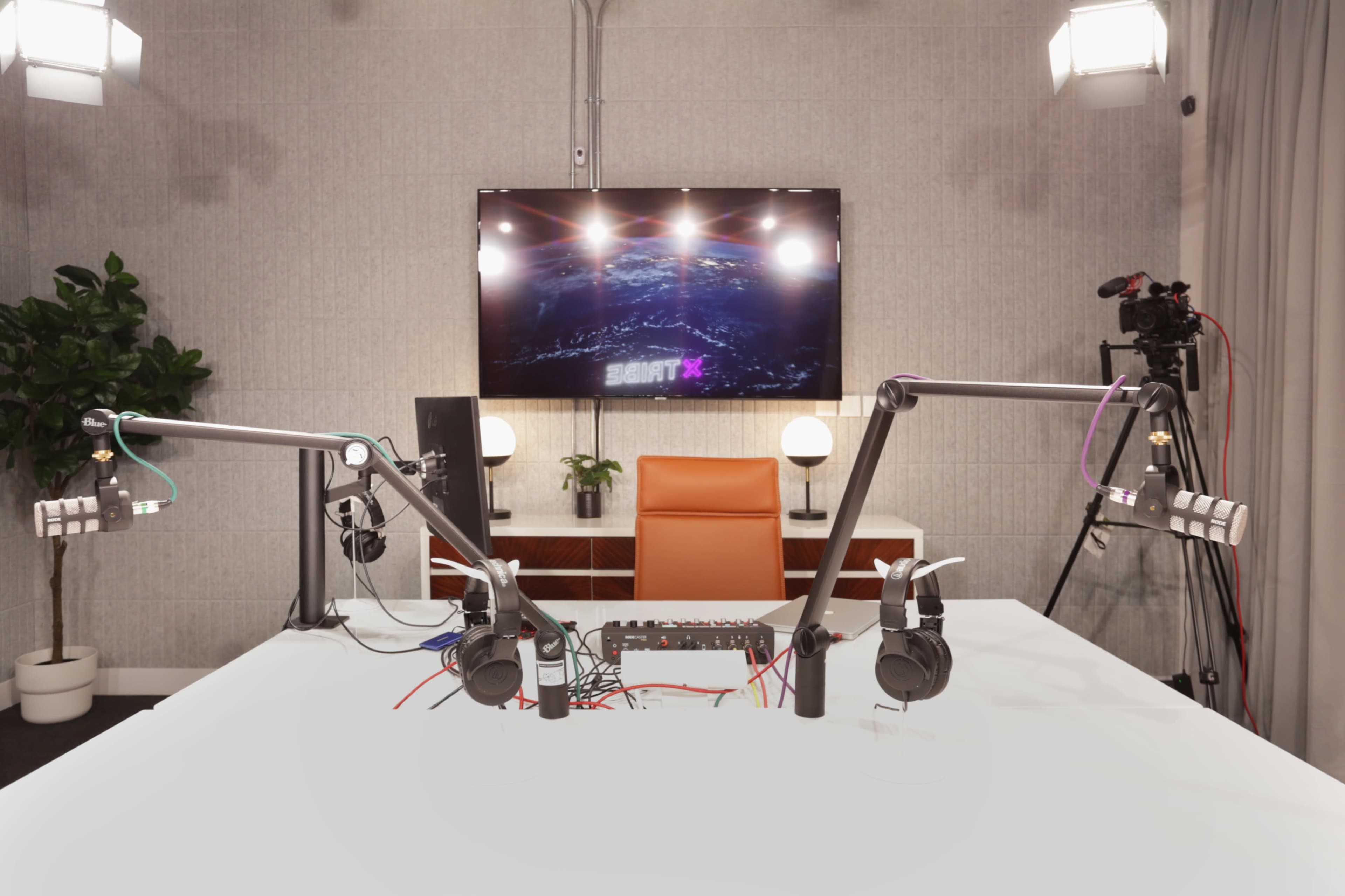 A podcast recording studio features two microphone setups, a central table with audio equipment, and a TV screen displaying a graphic.