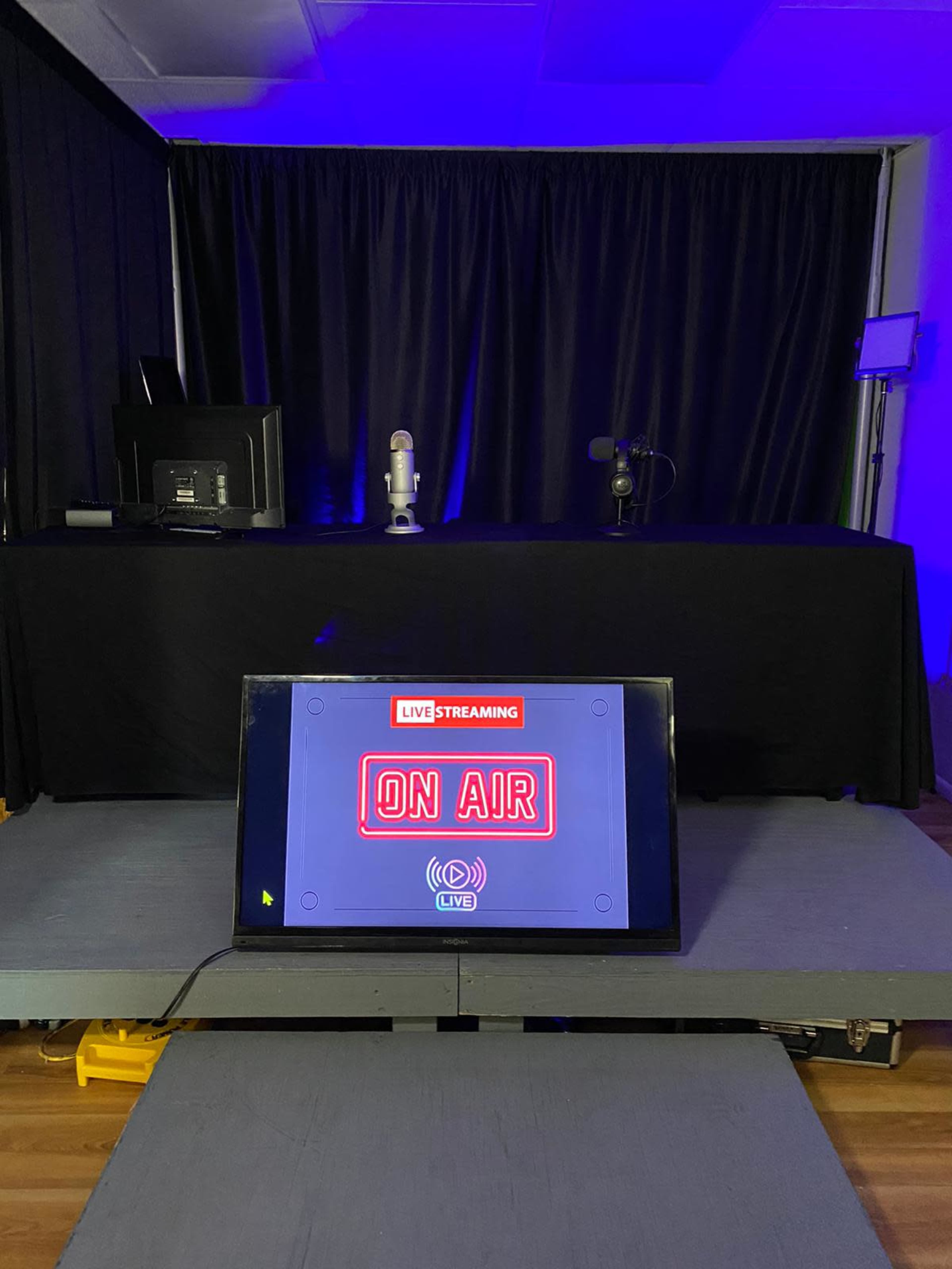 A studio setup features a microphone and computer behind a black table, with a lit "ON AIR" sign displayed on a monitor in the foreground.
