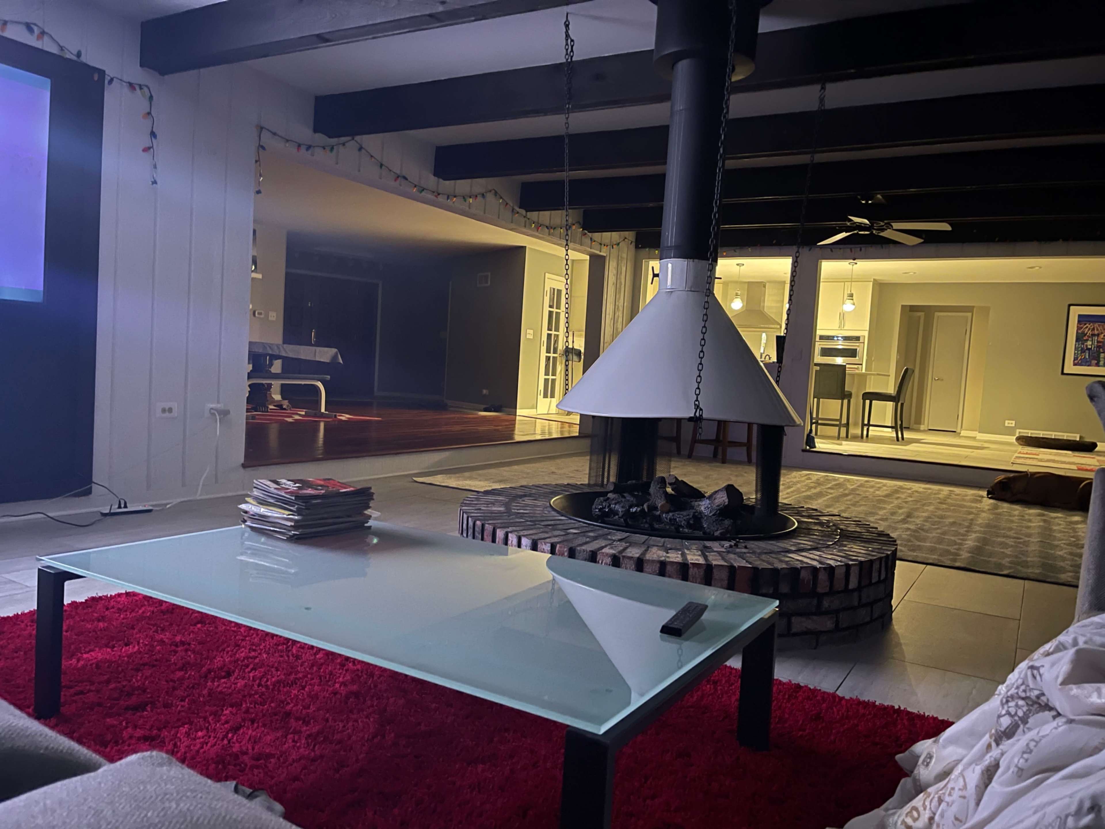 A spacious living area with a glass coffee table on a red rug, featuring a circular fireplace at the center and an open kitchen visible in the background.