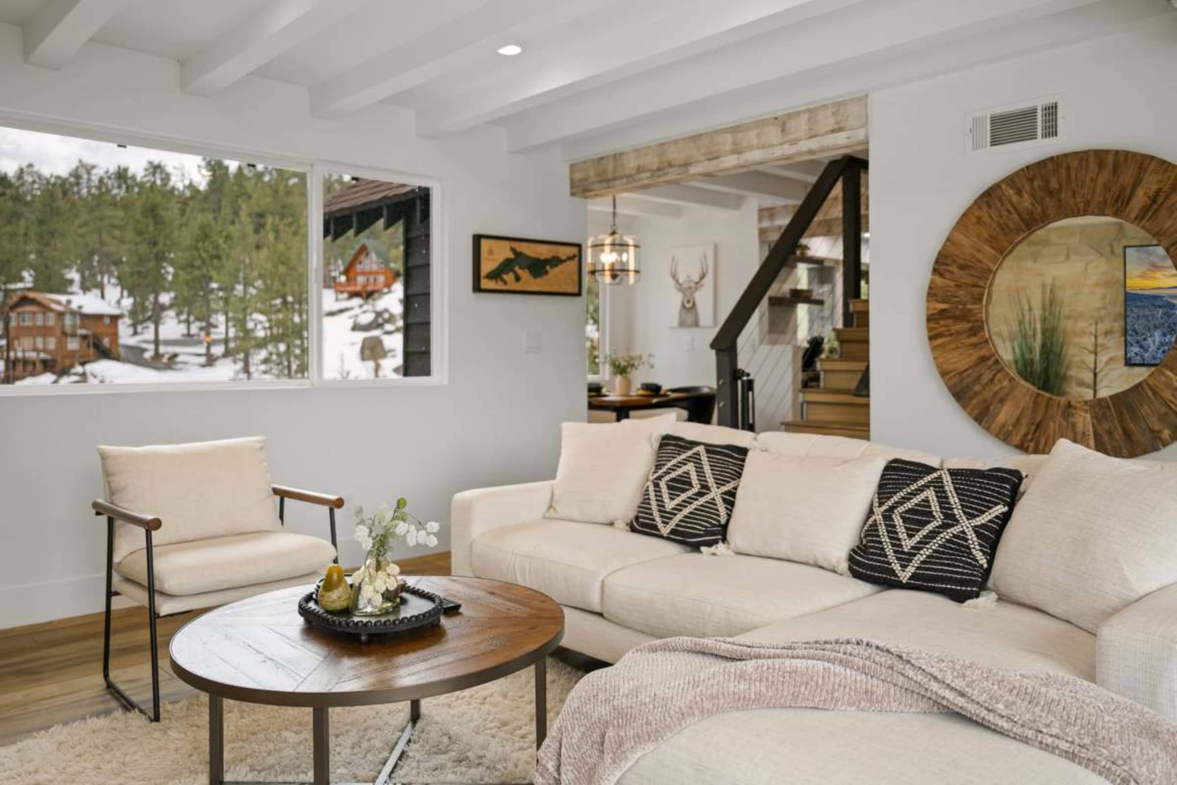 A cozy living room with a sectional sofa, a round wooden coffee table, and a large window offering a view of a snowy landscape and forested area outside.