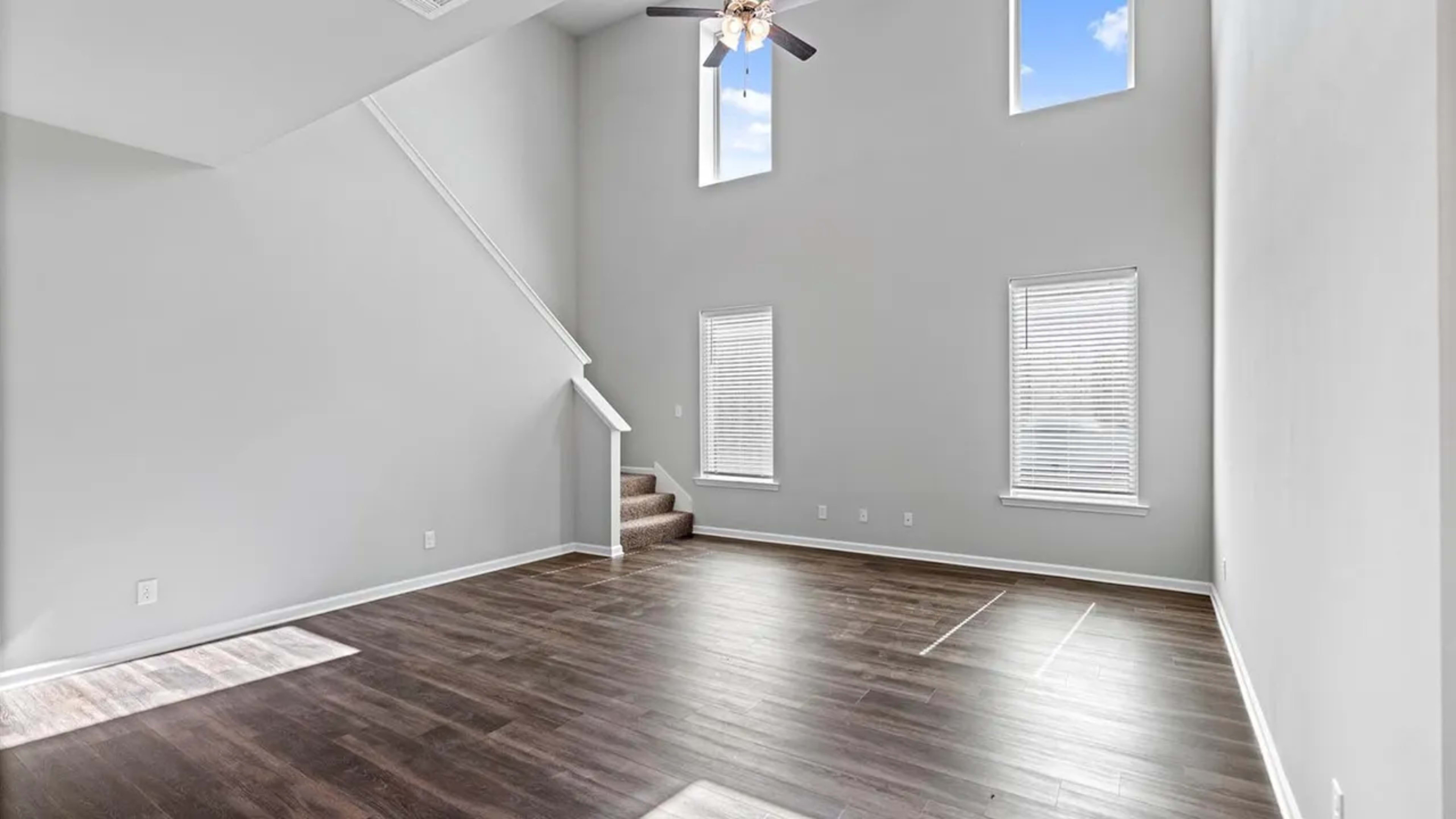A spacious living room with a ceiling fan, large windows, and a staircase leading to the upper floor.