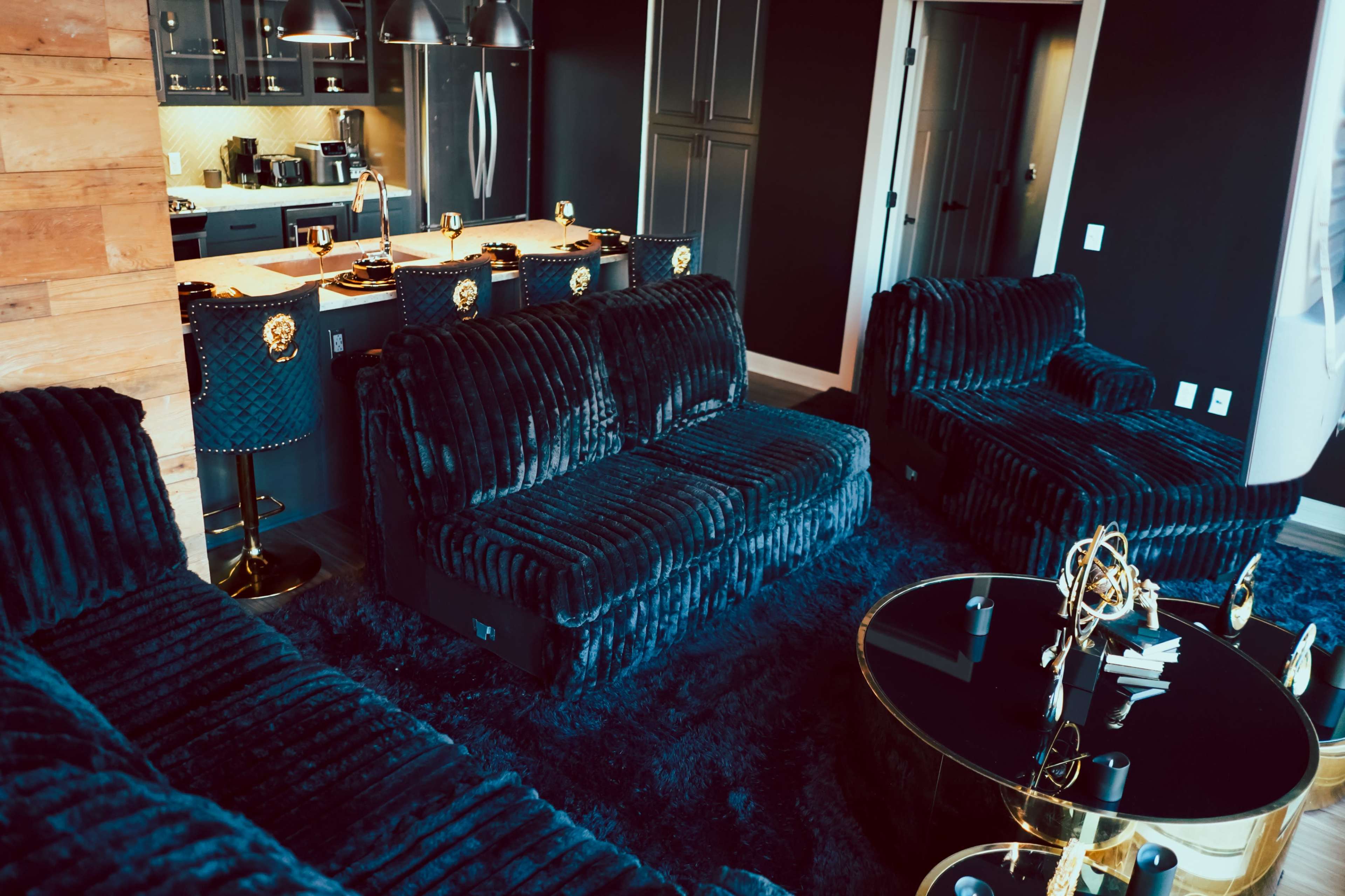 The image shows a stylish living area with dark, plush sofas, a glass coffee table, and a kitchen visible in the background.