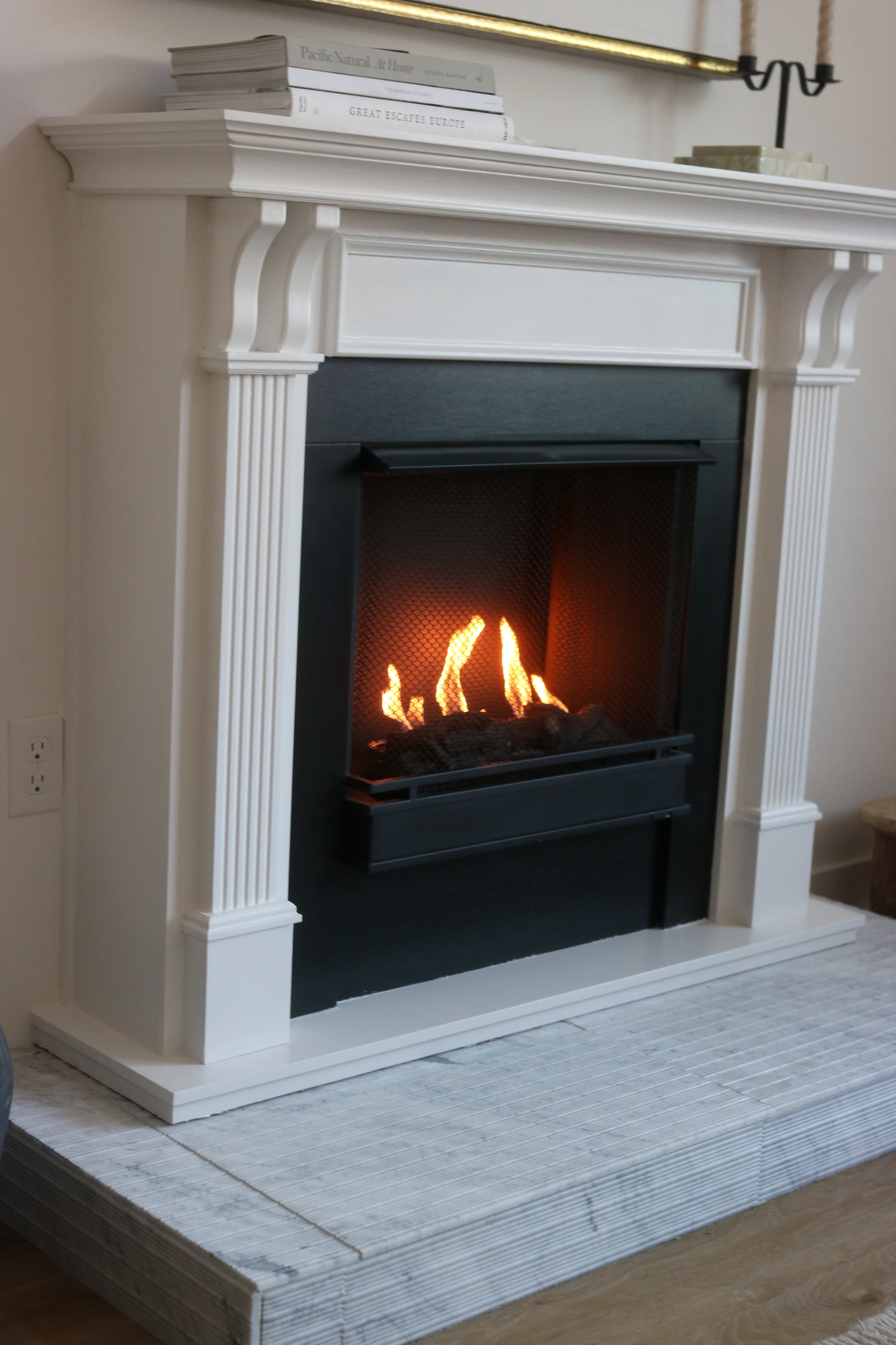 A modern fireplace with a black mesh front and flames visible is set into a white mantel with decorative columns, placed on a tiled hearth.