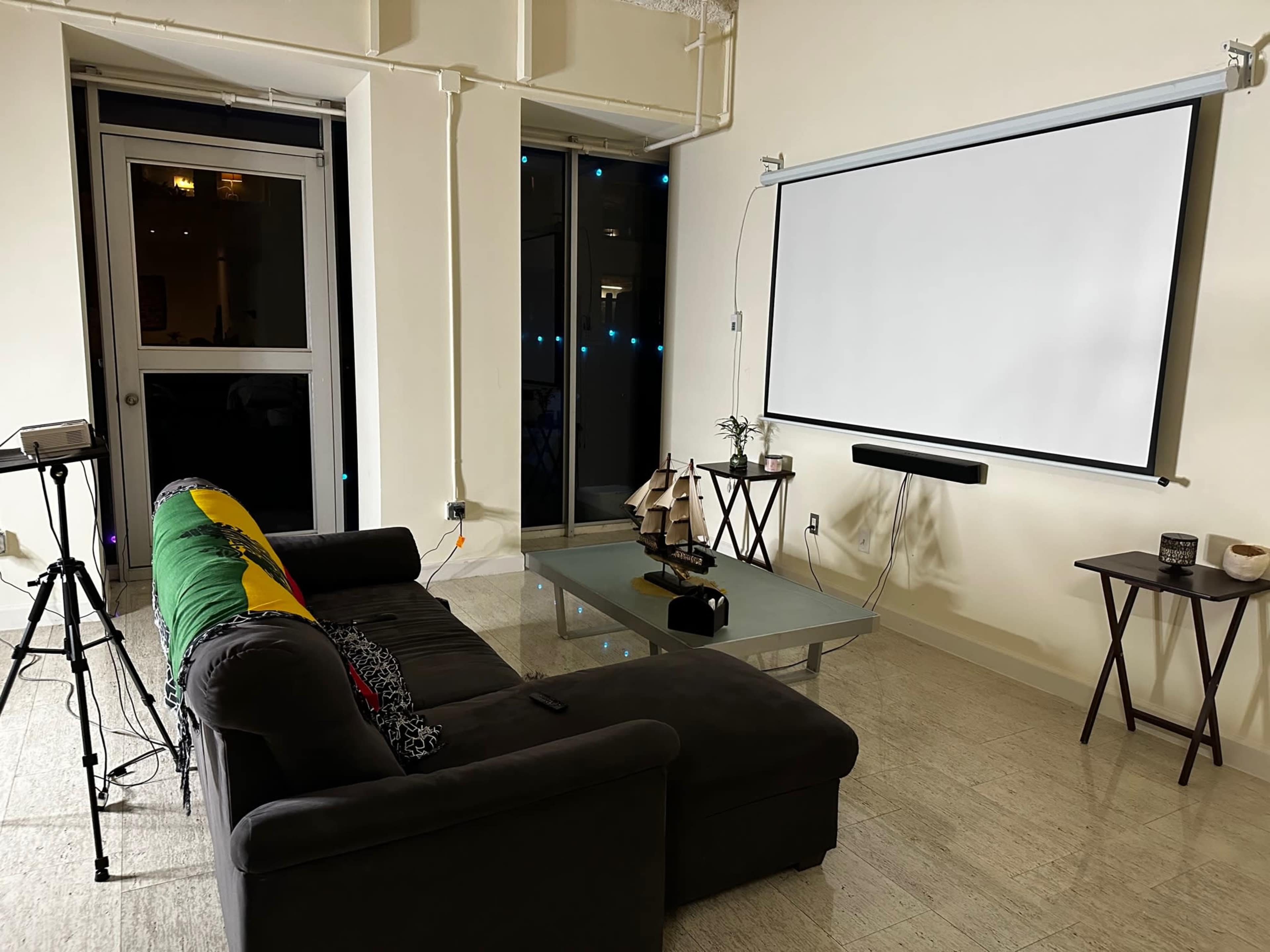 A modern living room features a dark sofa, a glass coffee table, a wall-mounted projector screen, and a small table with decorative items.