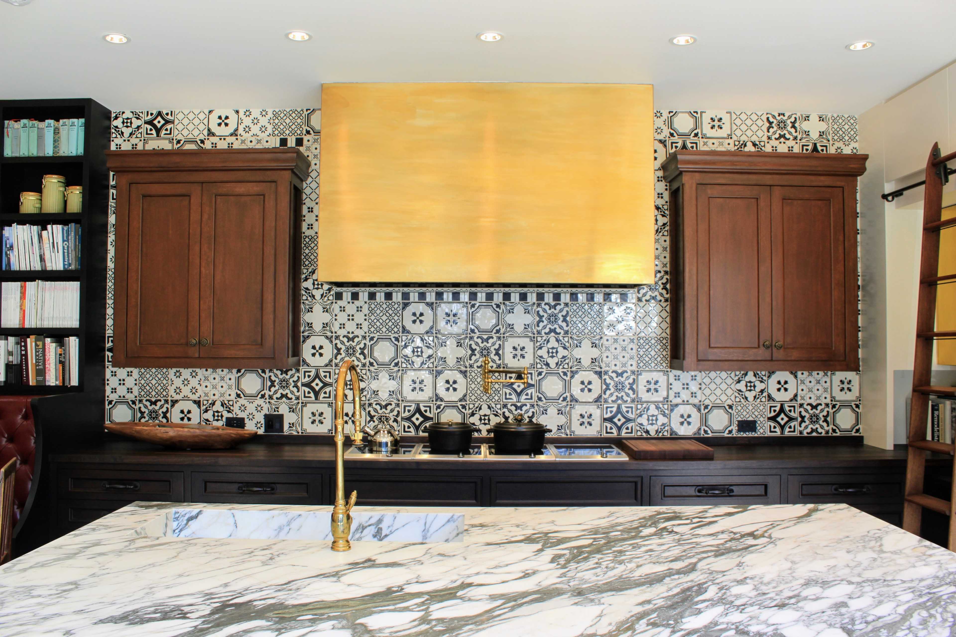 A kitchen with a marble countertop, wooden cabinets, and a decorative tiled backsplash behind a gold-colored range hood.