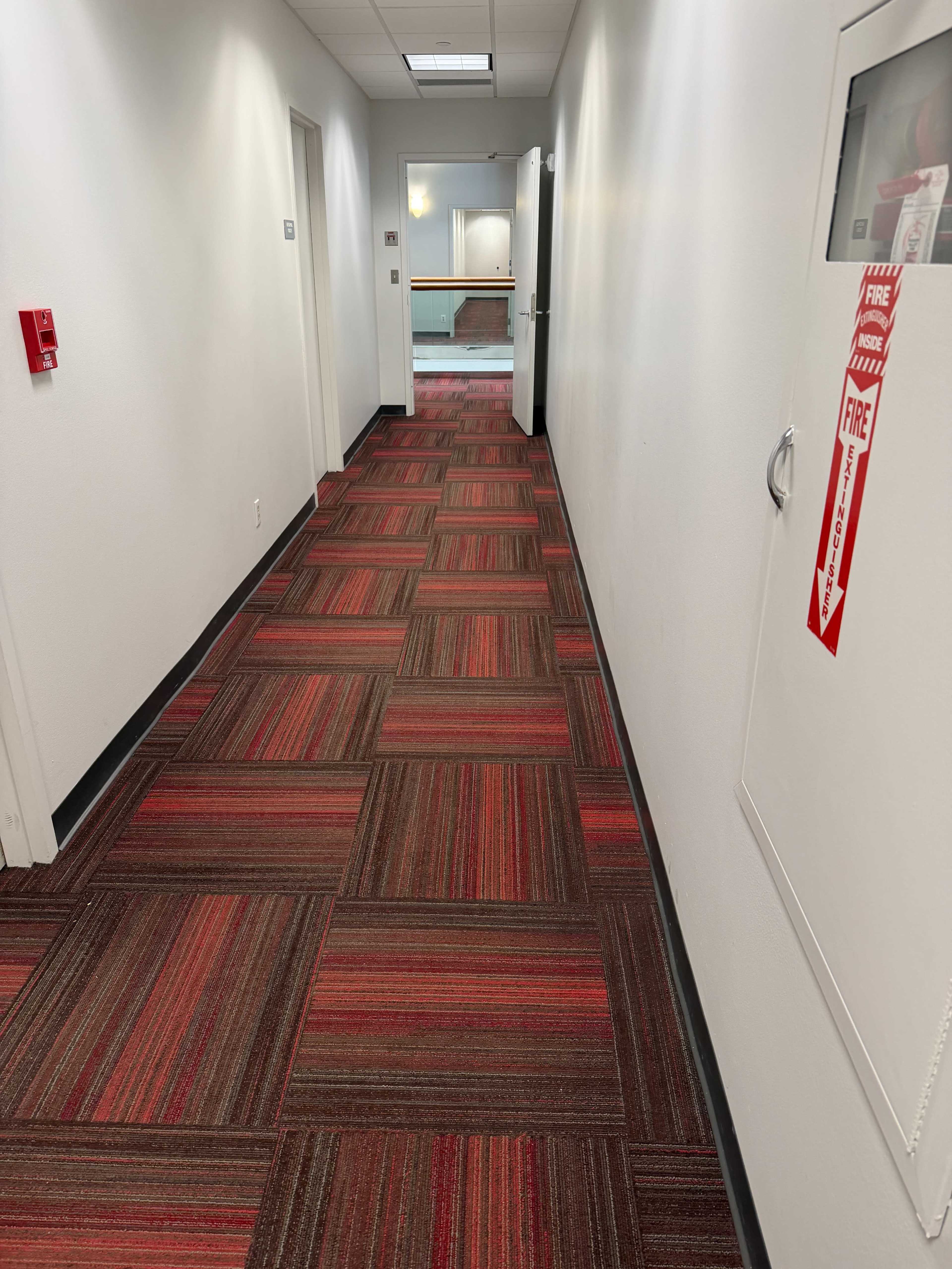 A long hallway features red and gray patterned carpet, with doors on either side and a fire extinguisher station on the right wall.