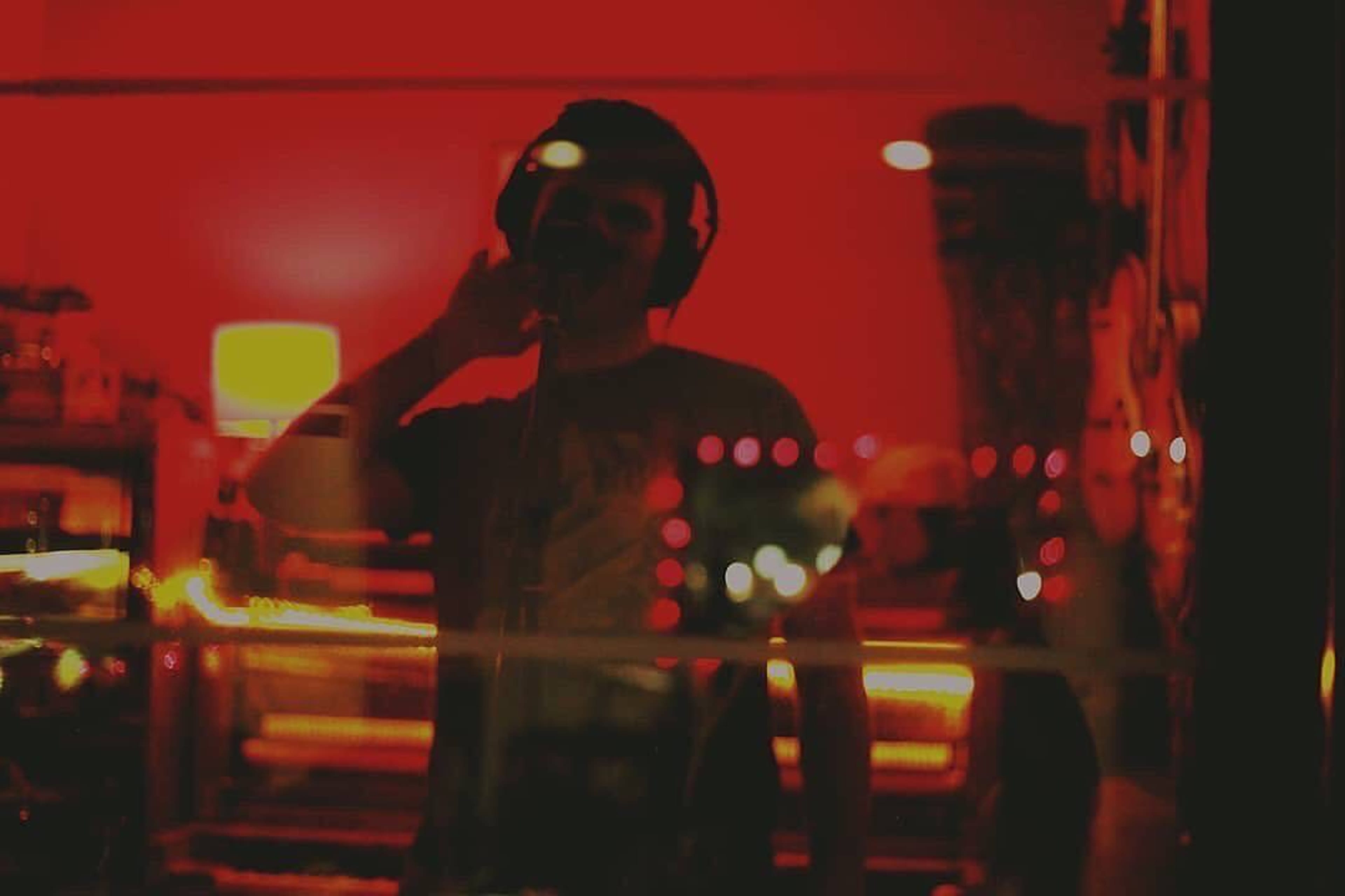 A person wearing headphones stands in front of a microphone, illuminated by red lighting, with reflections from surrounding lights visible in a window.