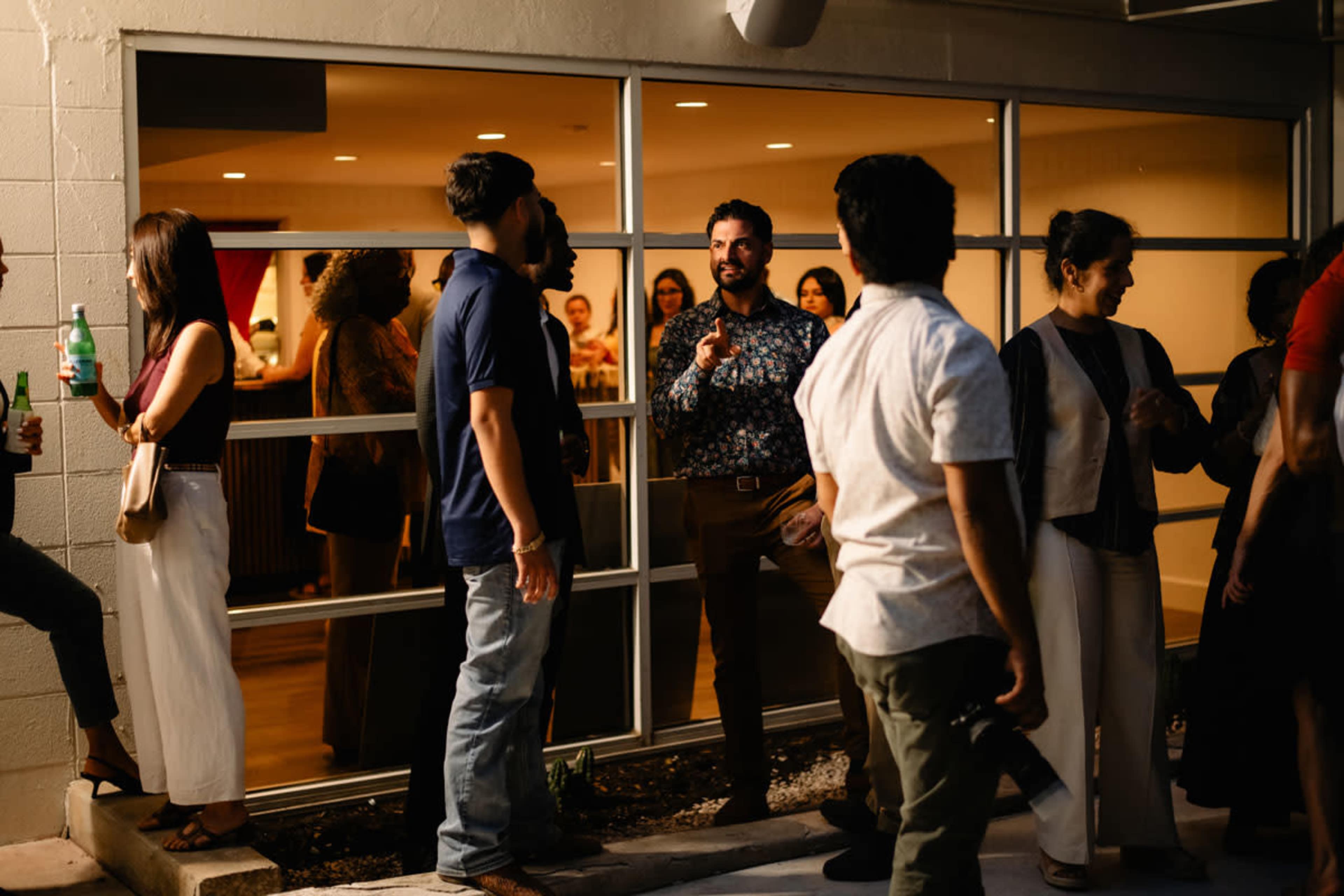 A group of people socialize outside a large window, with some individuals holding drinks and engaging in conversation.