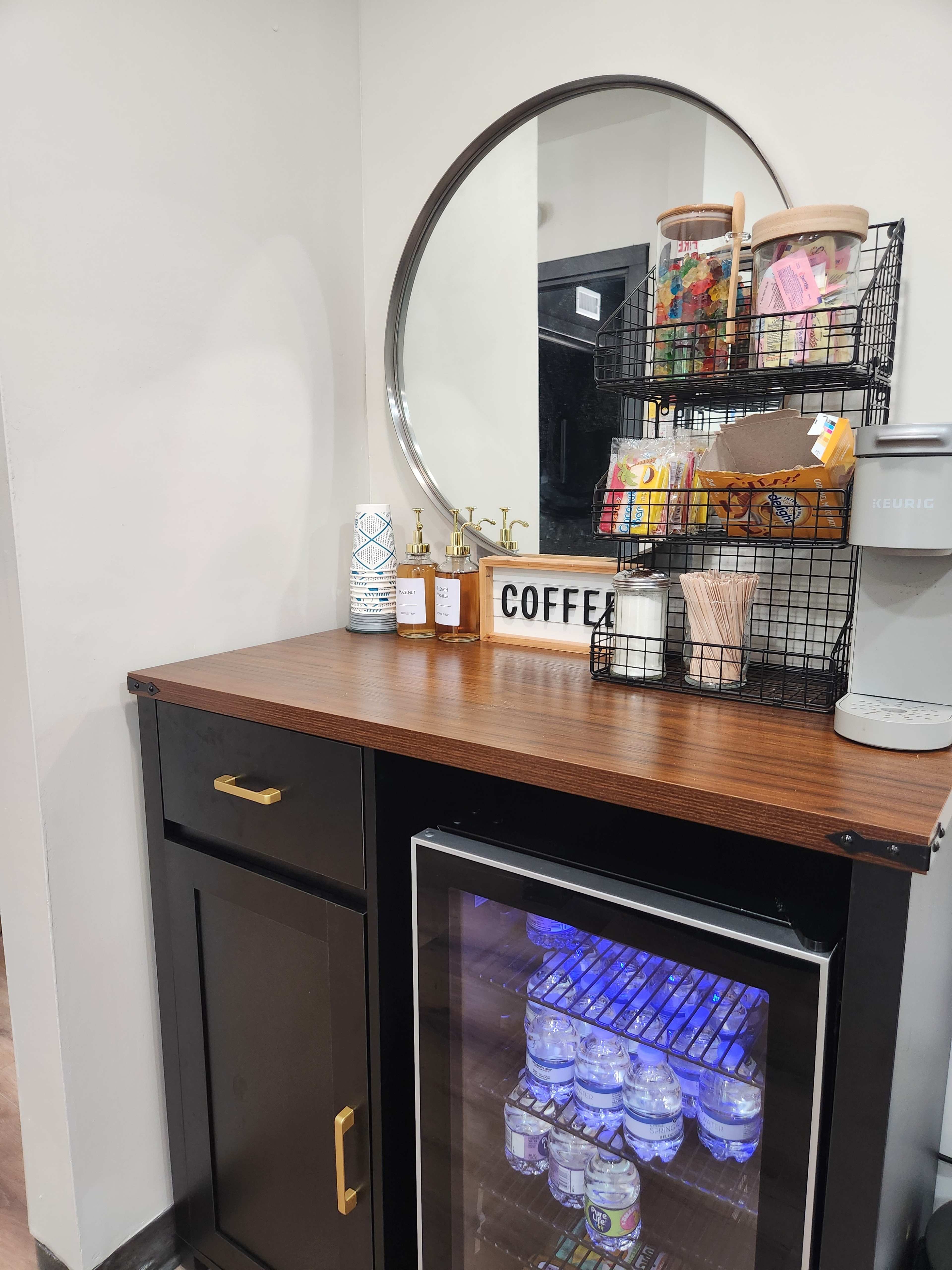 The image shows a compact coffee station featuring a wooden countertop, a circular mirror, a selection of coffee supplies in a wire basket, decorative items, and a mini fridge displaying bottled water.