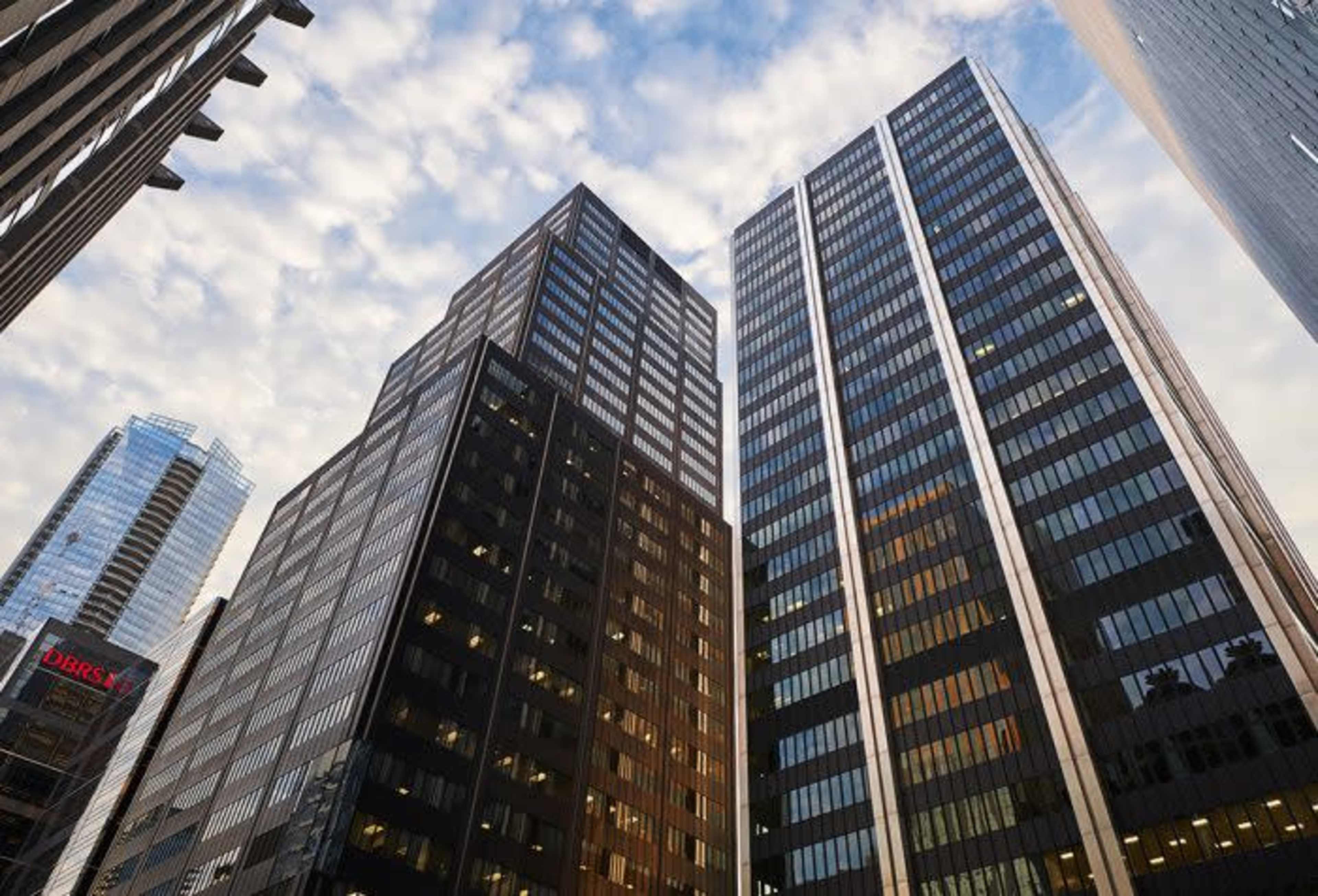Tall office buildings with glass facades rise against a cloudy sky in an urban environment.