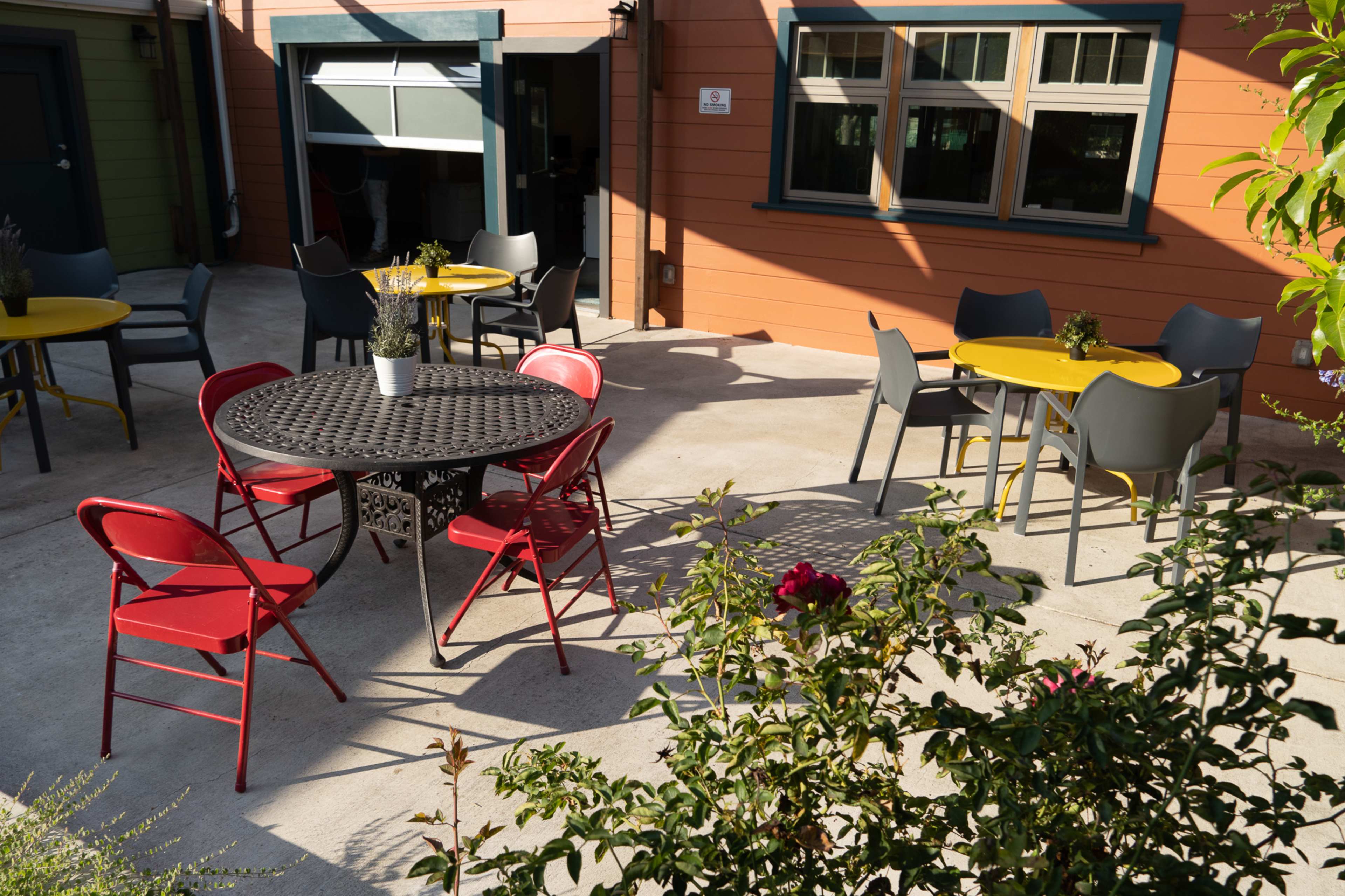 A small courtyard features a round black metal table surrounded by red and gray chairs, with several yellow-topped tables positioned nearby and greenery in the foreground.