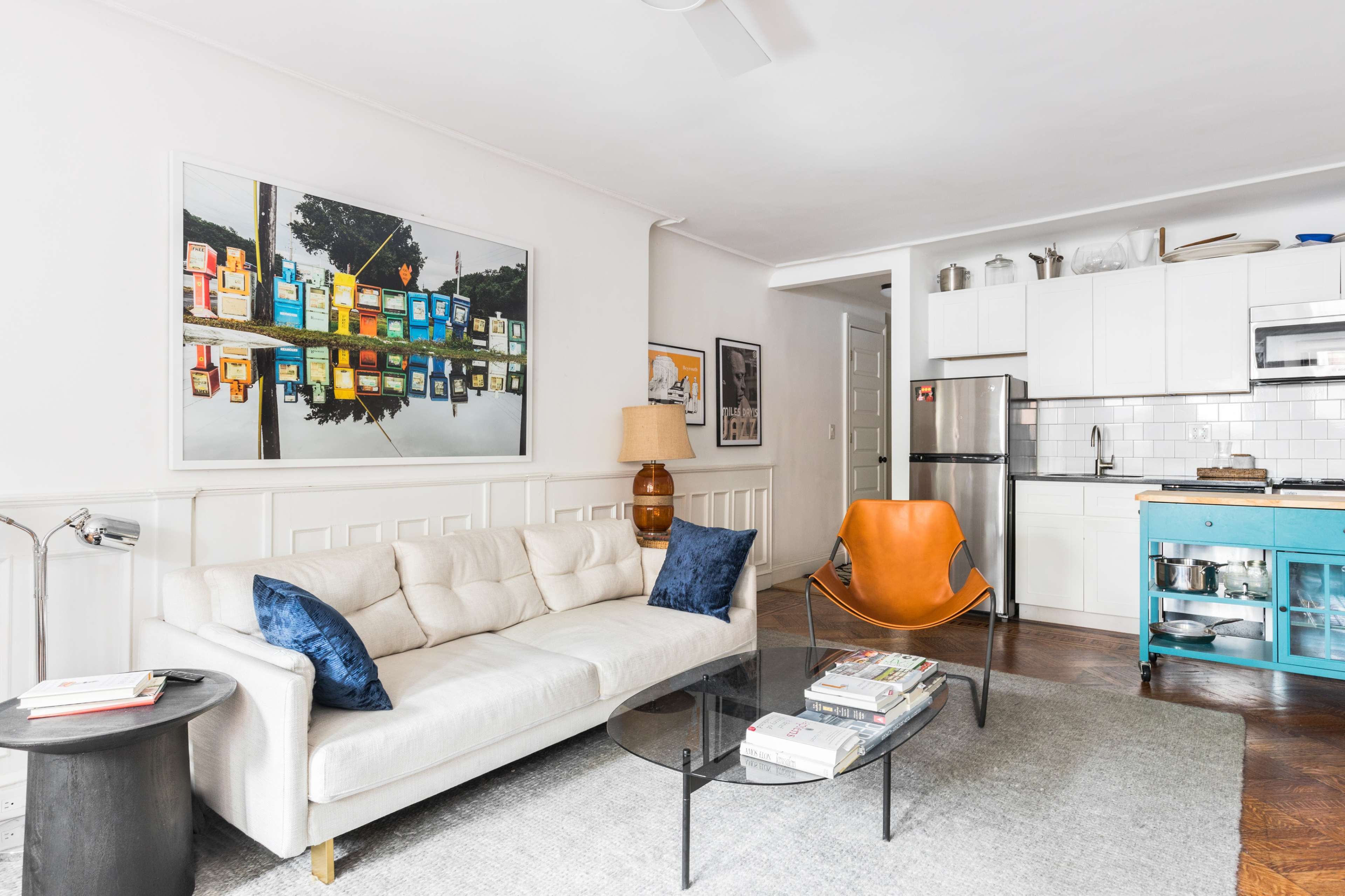 The living area features a white couch, a glass coffee table, an orange chair, and a kitchen with white cabinetry in the background.