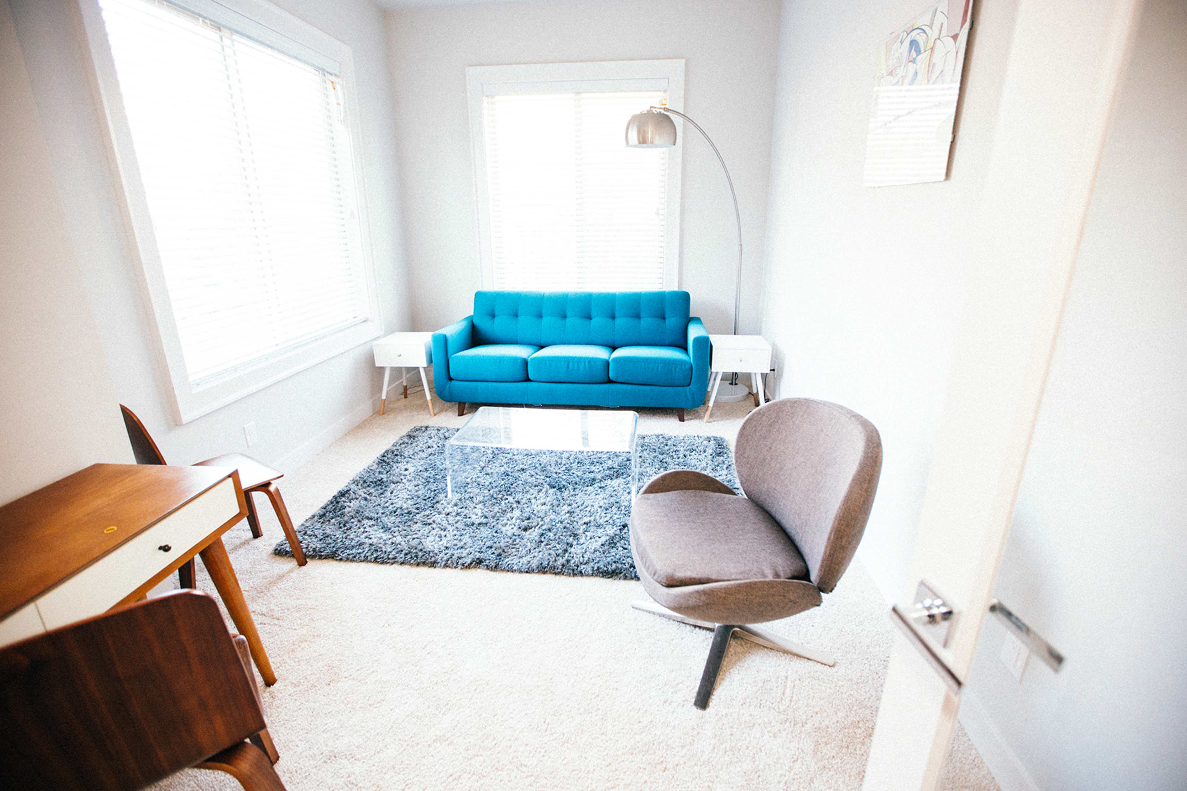 A small, modern living room features a blue couch, gray chair, glass coffee table, and a light rug on carpet.