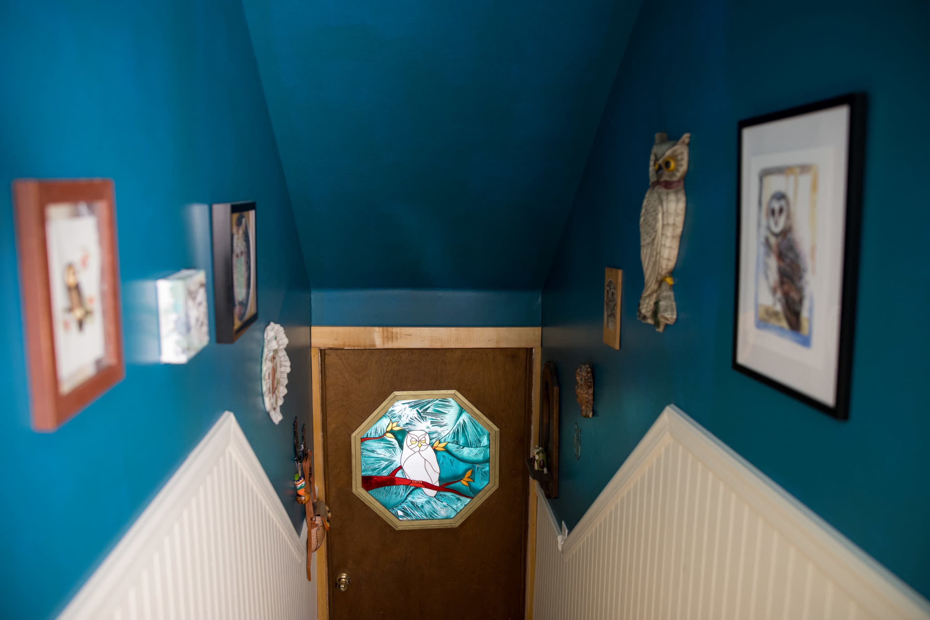 The image shows a narrow staircase leading to a door with a colorful octagonal stained glass window, surrounded by framed artwork on the teal walls.
