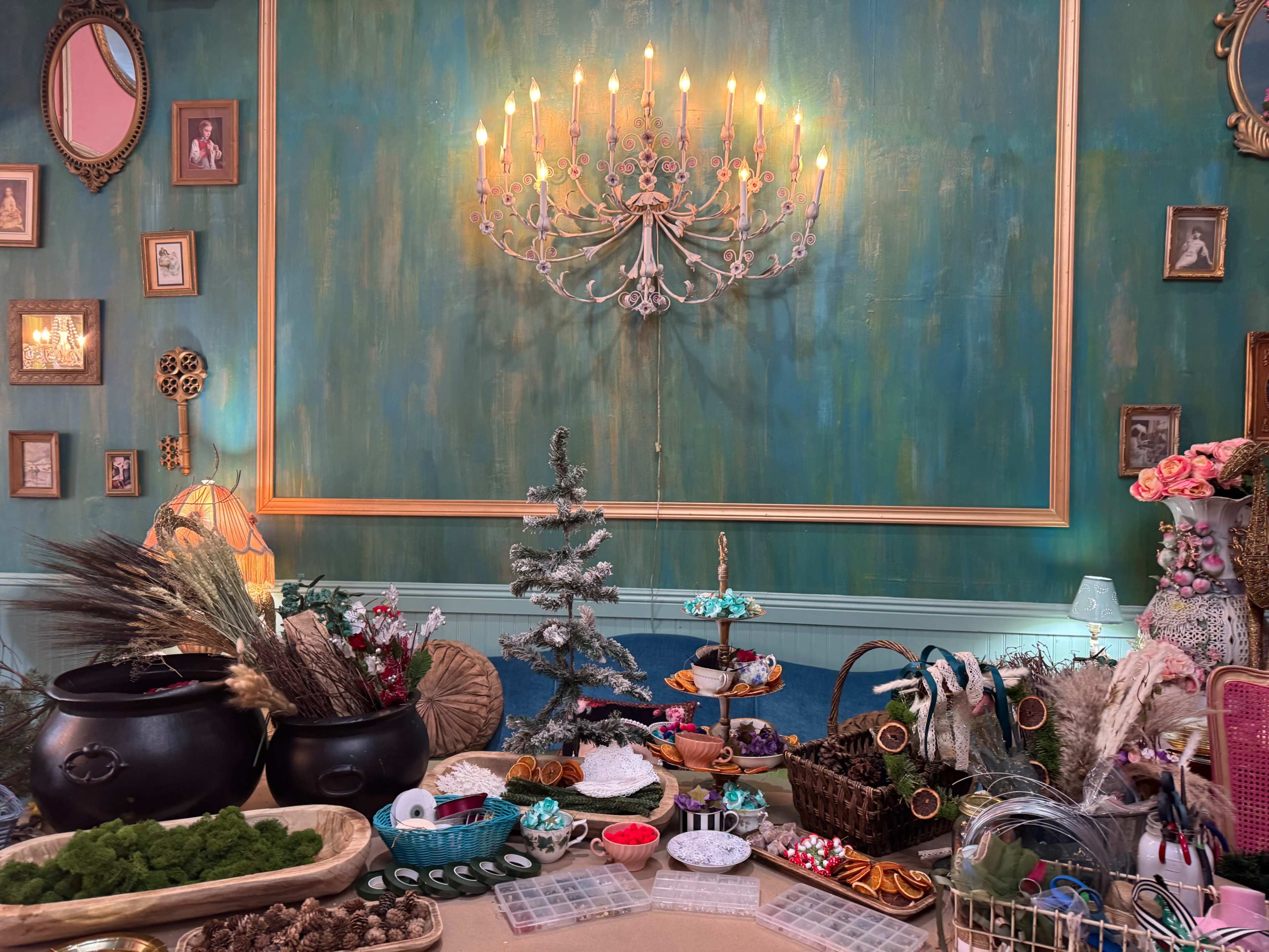 The scene features a decorated table with various holiday-themed items, including a small Christmas tree, decorative plants, and an assortment of foods and crafts against a vibrant green wall with framed pictures.