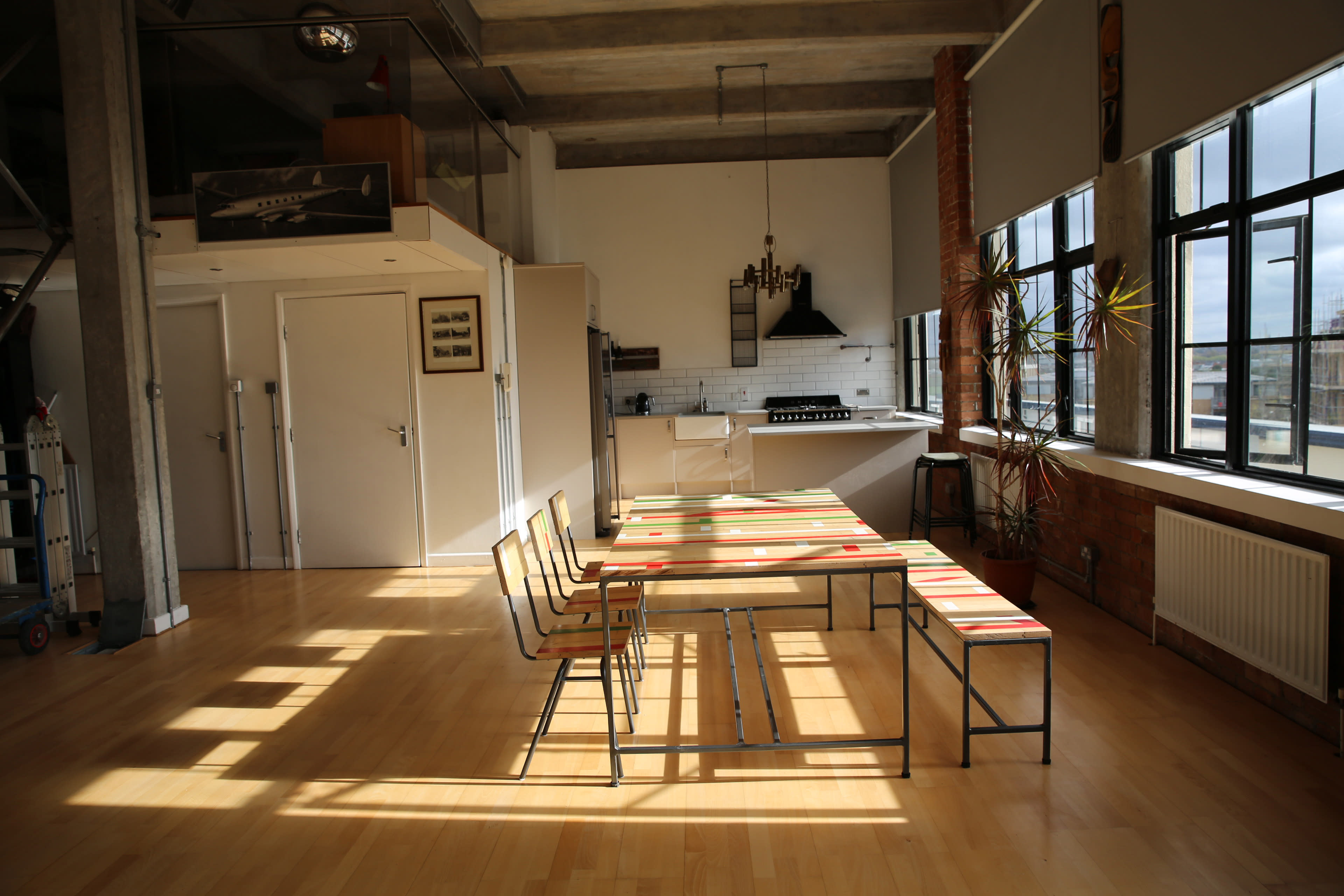The image shows a spacious open-concept dining area with a long table and several chairs, illuminated by natural light from large windows.