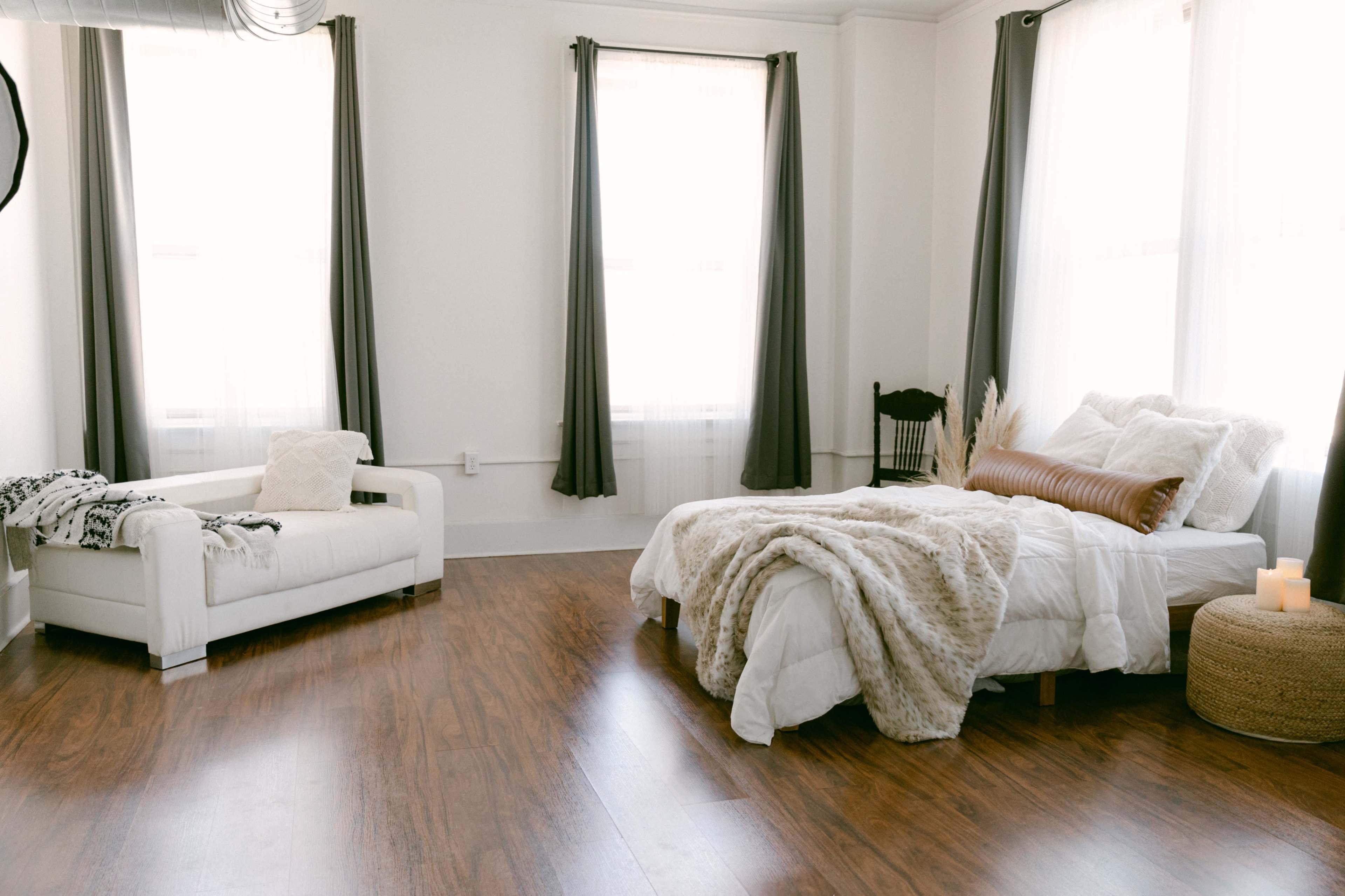 A bright and airy room with a bed dressed in white linens and a cozy throw, a white couch, and large windows dressed with grey curtains.
