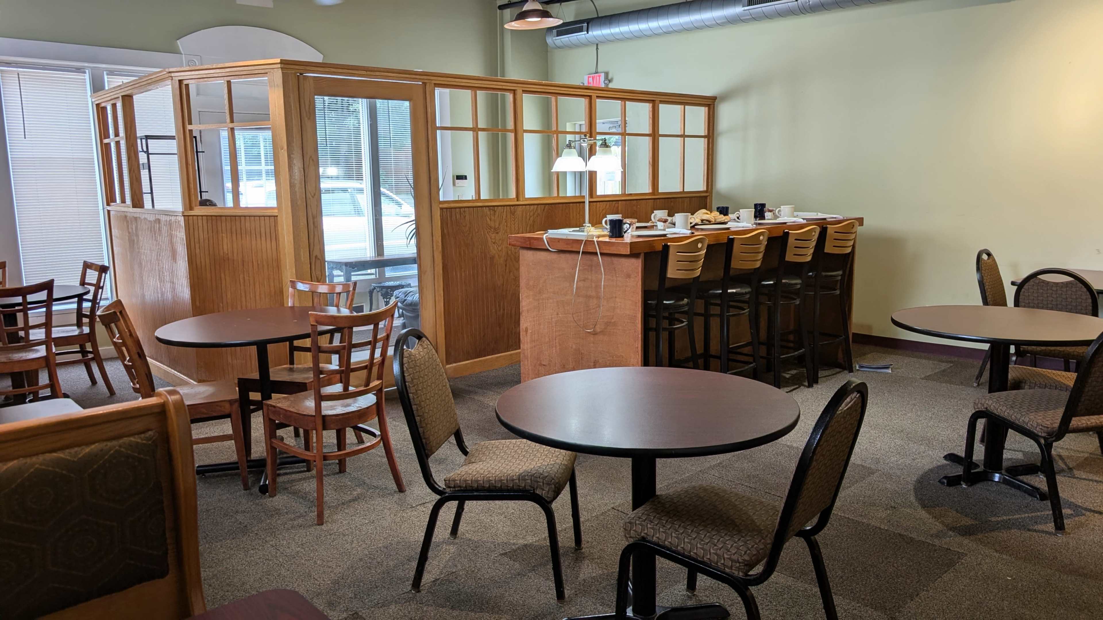 The image shows a café interior with a seating area featuring round tables and chairs, and a bar counter behind a wooden partition.