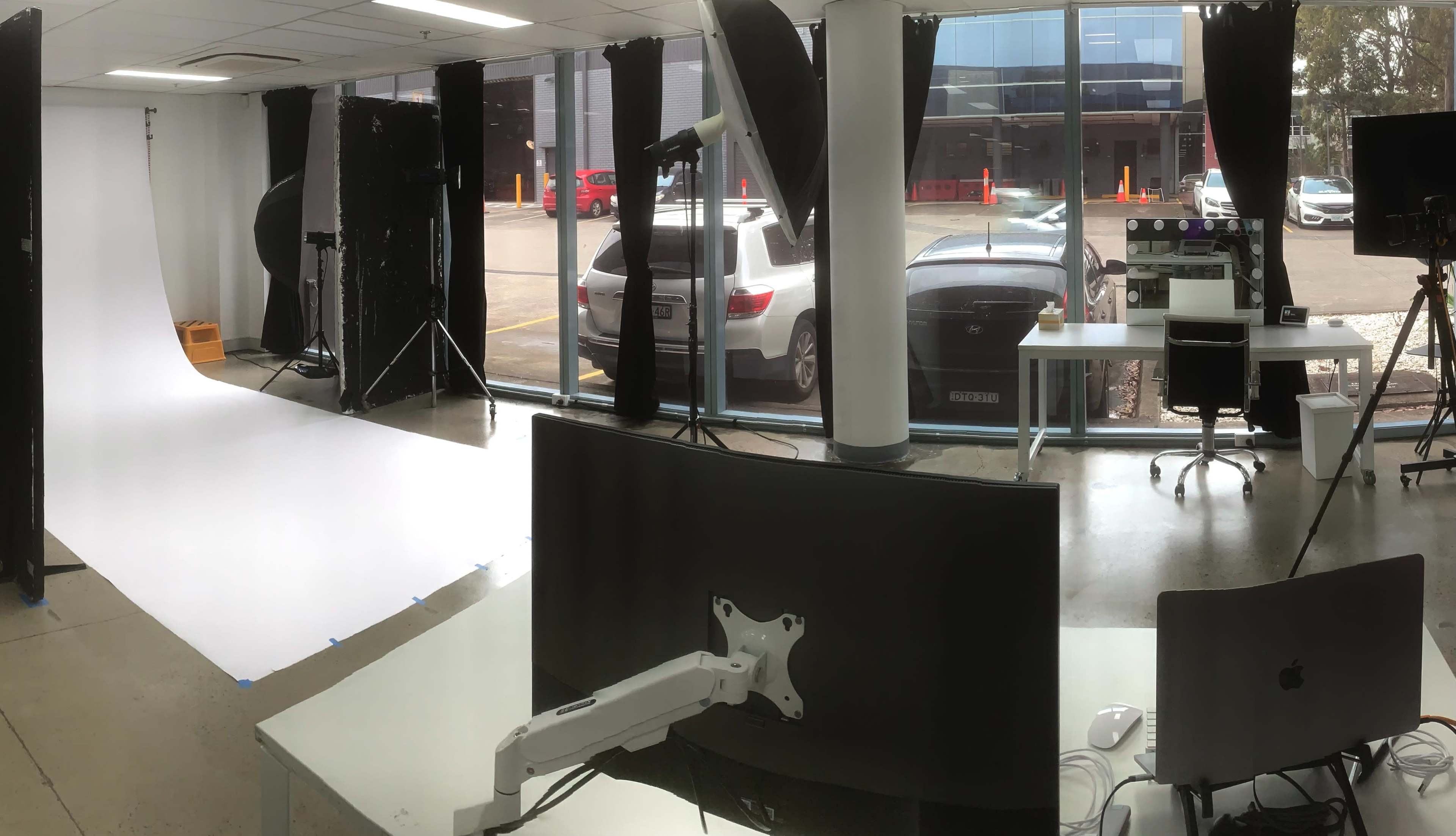A photography studio with a white backdrop, lighting equipment, and a desk facing a large window overlooking parked cars.