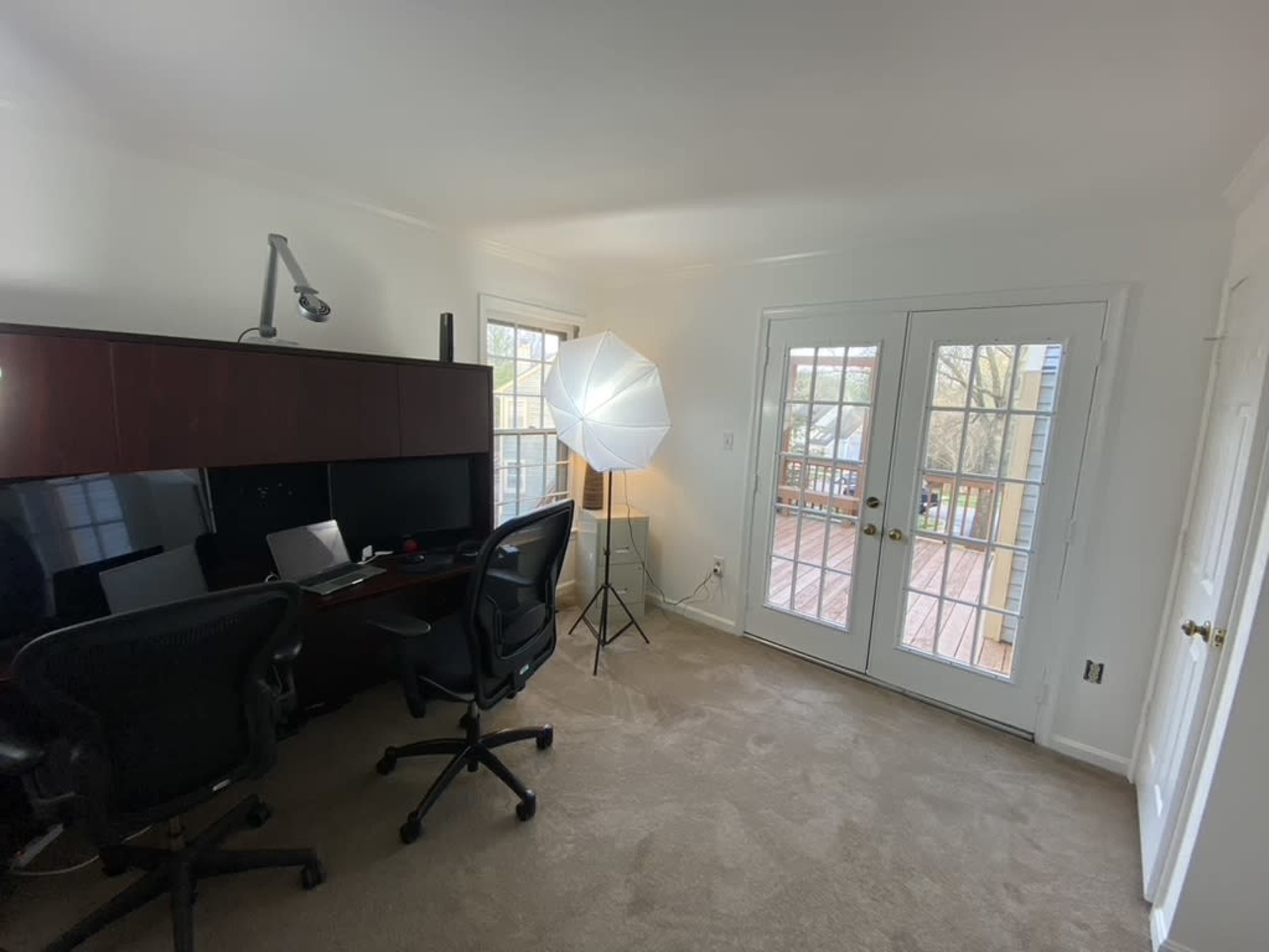 A well-lit office space with a wooden desk, two office chairs, and a softbox lighting setup next to a set of French doors leading to a deck.
