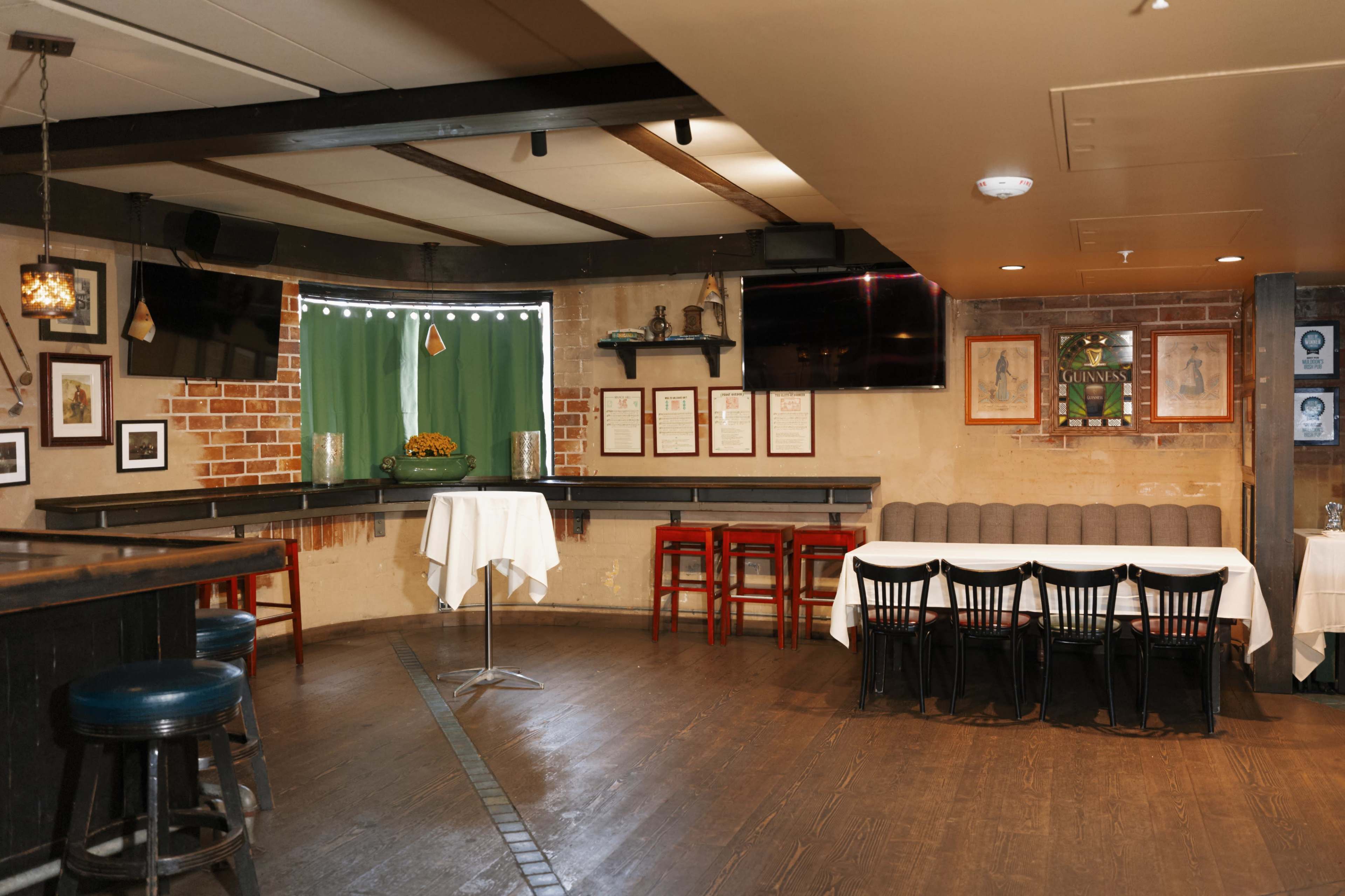 The interior of a restaurant features a mix of seating options, including a high table, bar stools, and a long row of chairs along a wall, with exposed brick and two large screens mounted on the walls.