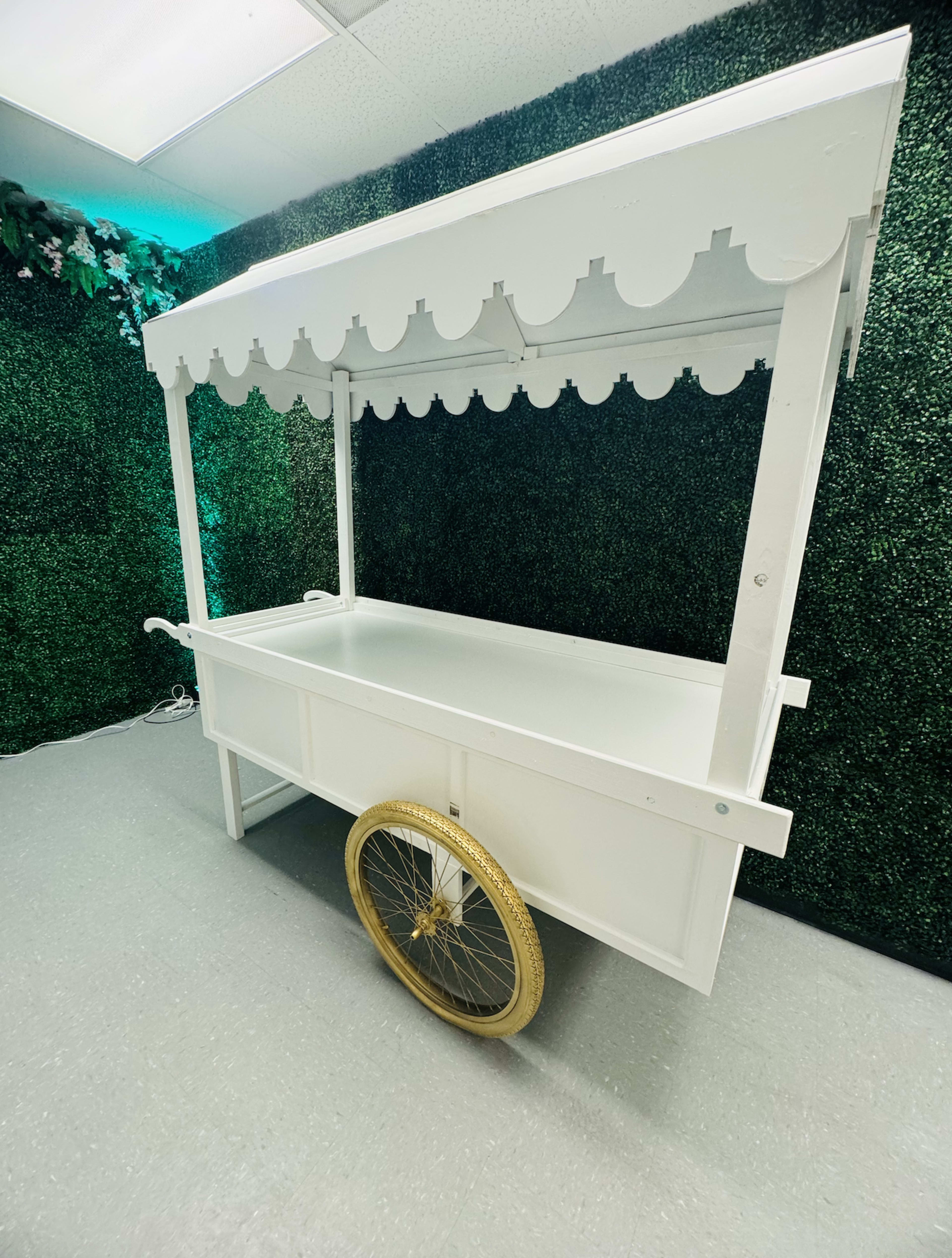 A white decorative cart with a scalloped roof and a gold wheel is situated against a green hedge backdrop.