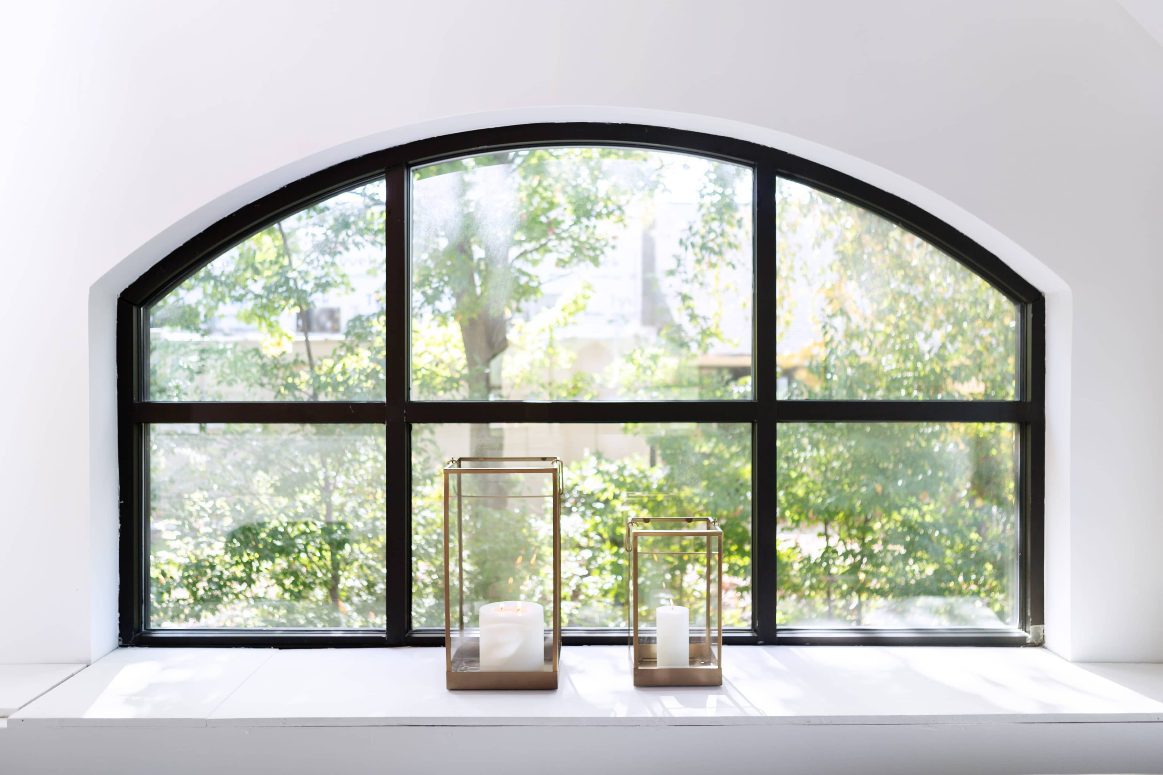 A large arched window frames two glass candle holders on a ledge, with greenery visible outside.