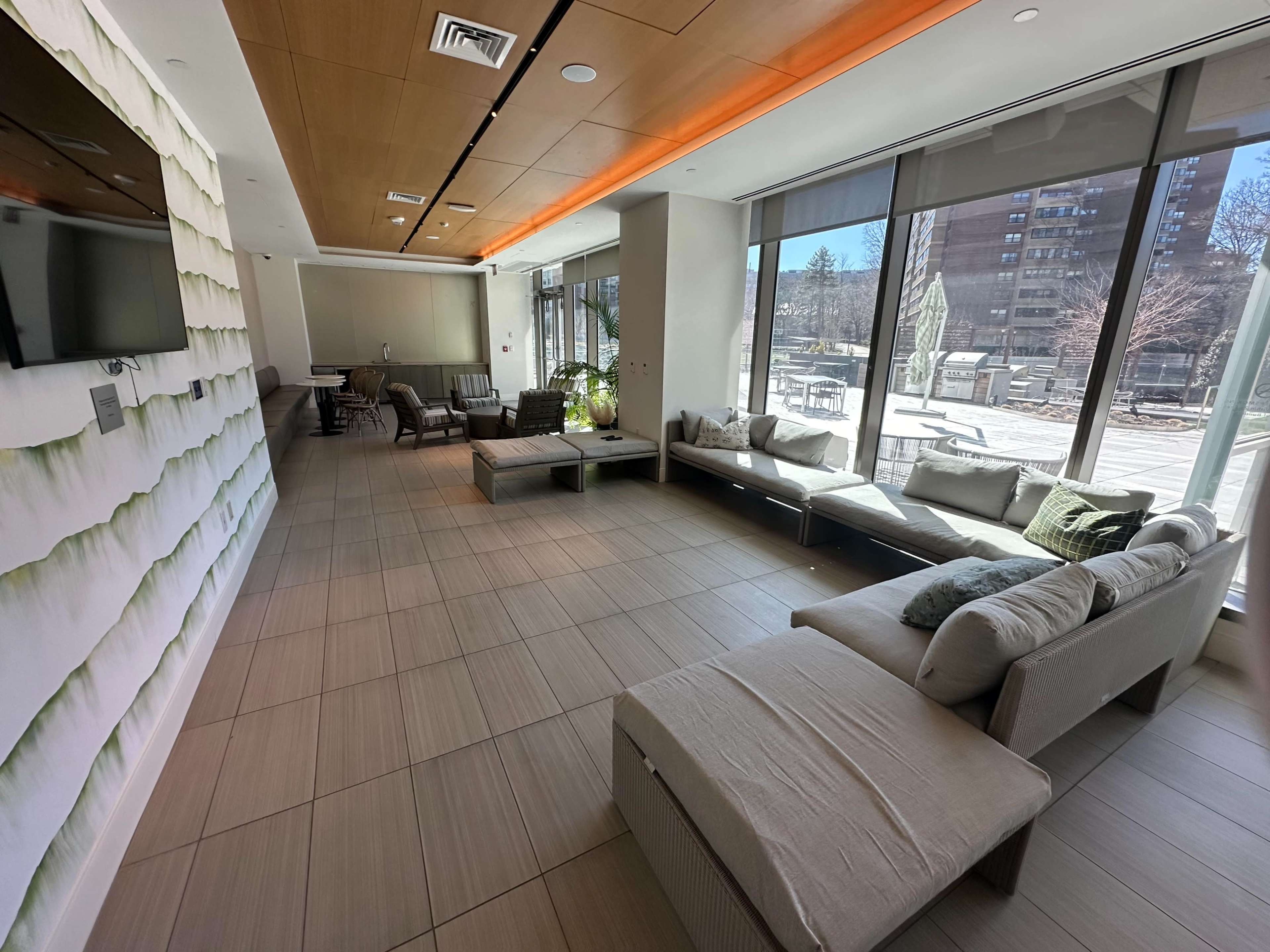 The image shows a spacious indoor lounge area with large windows, modern seating, and a TV on the wall.