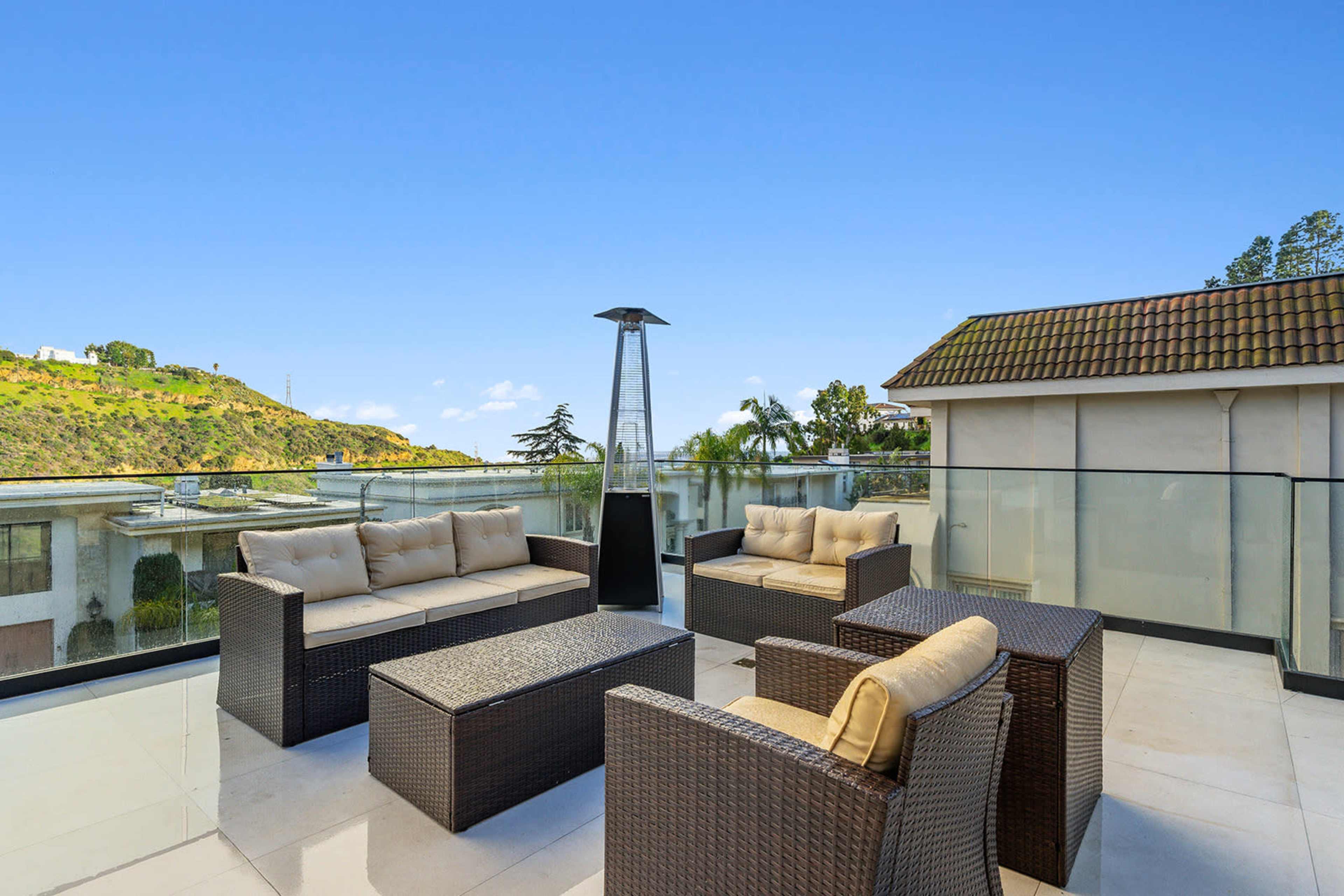 A terrace with outdoor seating, a glass railing, and a heater, overlooking a hilly landscape.