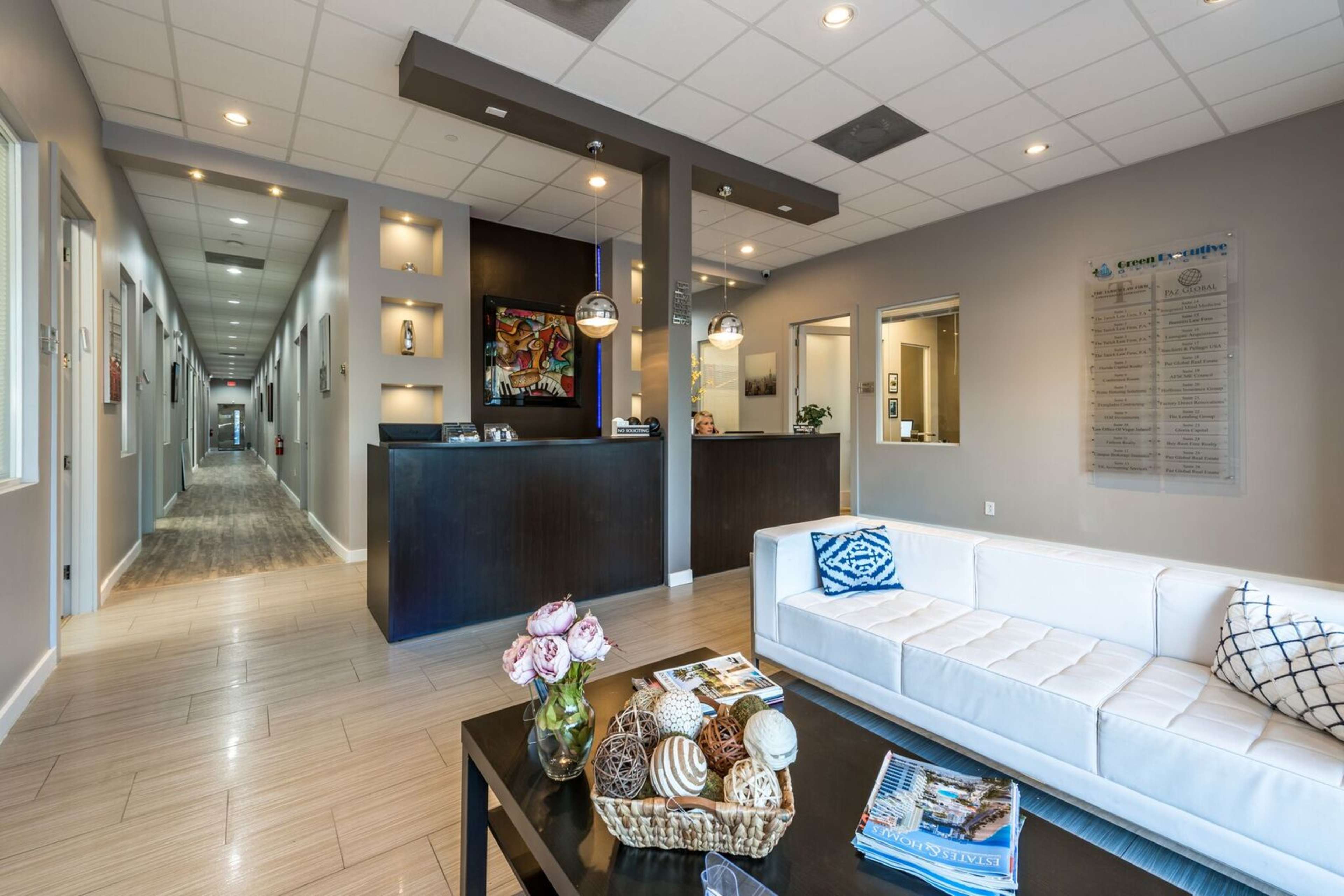 The image shows a modern reception area featuring a white couch, a coffee table with decorative items, and a front desk, with a corridor leading to multiple rooms.