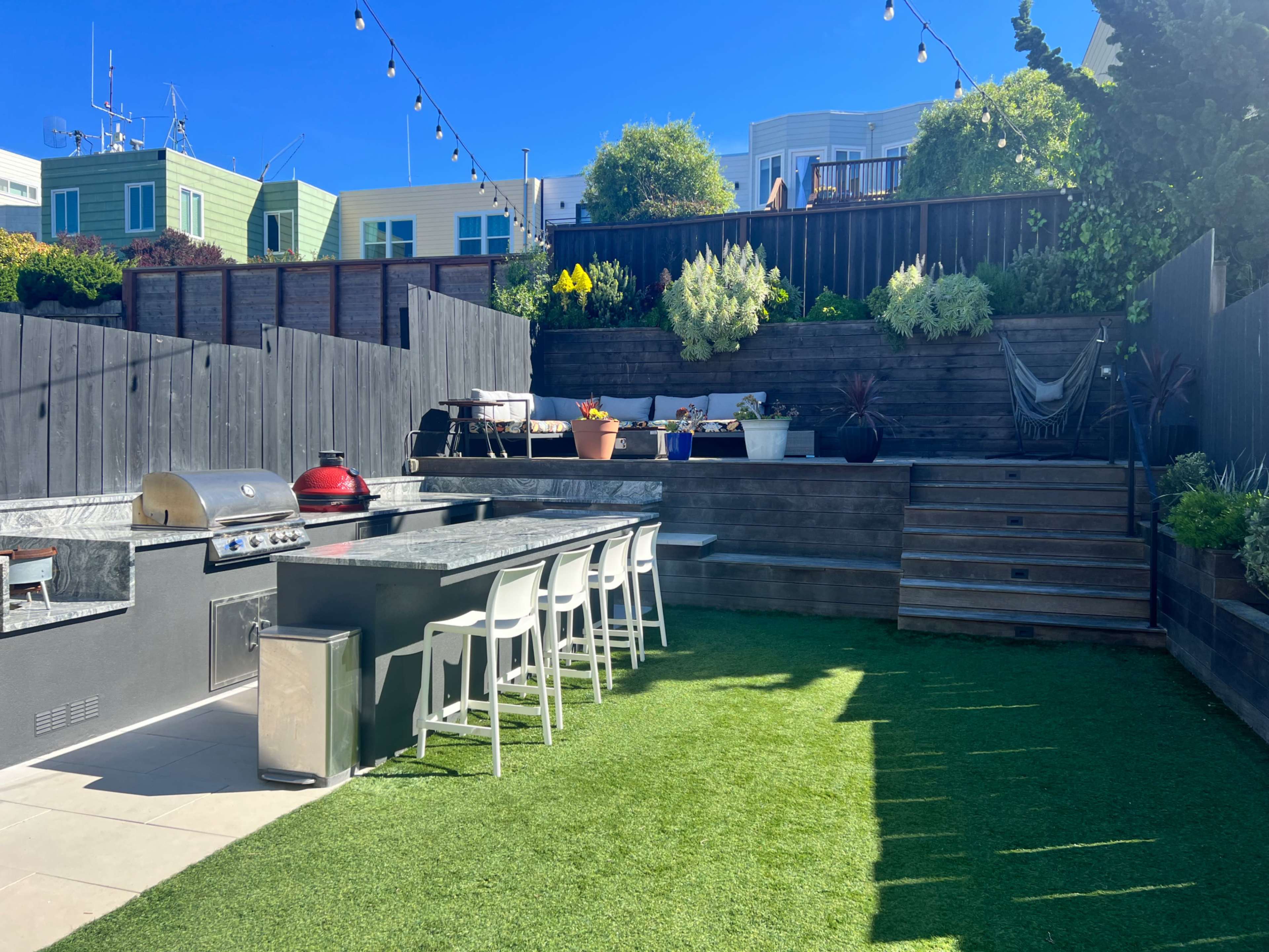 The image shows an outdoor kitchen area with a grill and seating on artificial grass in a multi-level garden surrounded by wooden fencing and various plants.